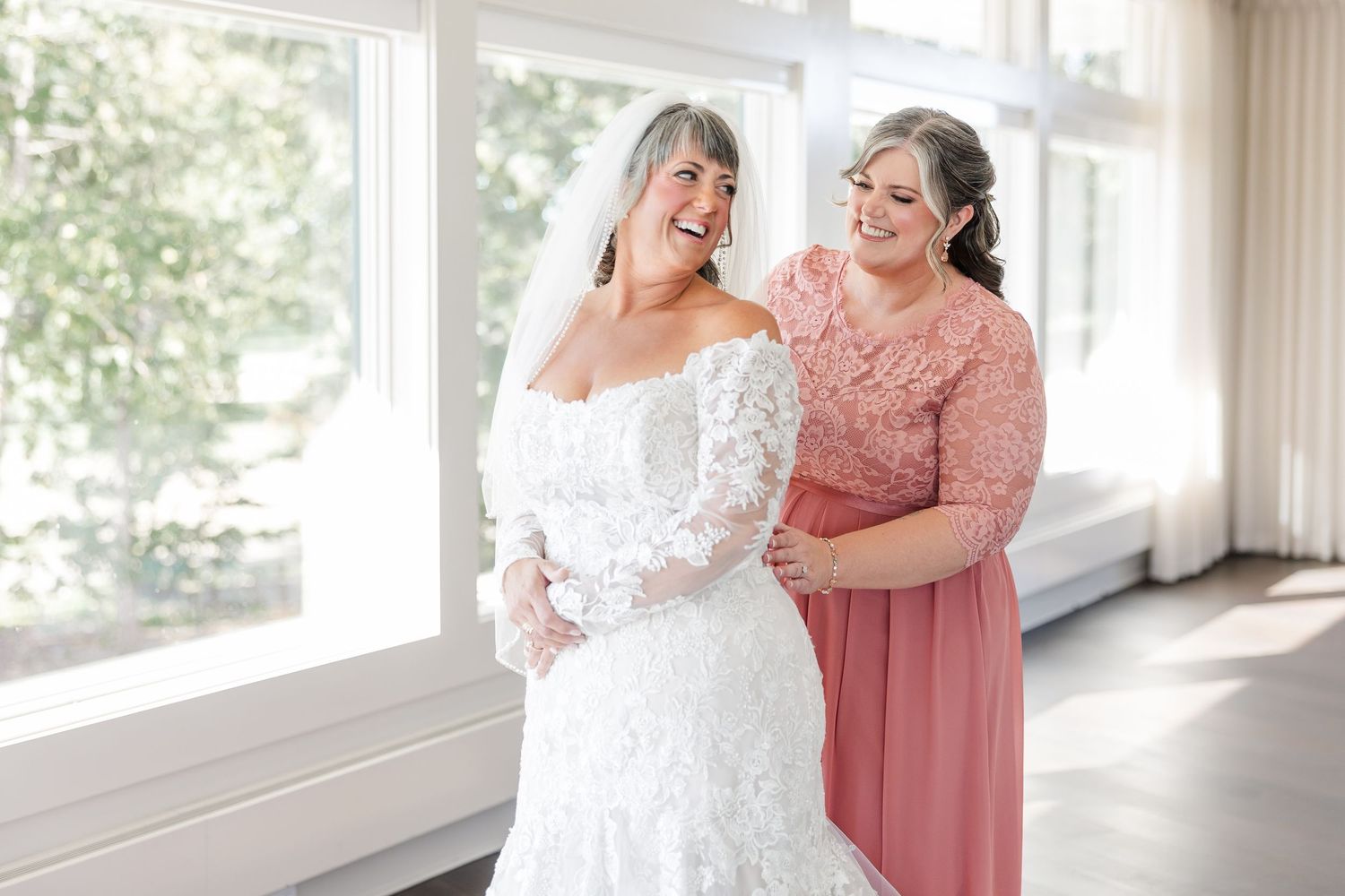 Smiling bride in a white lace wedding gown and veil shares a joyful moment with a woman in a coral dress near sunlit windows.