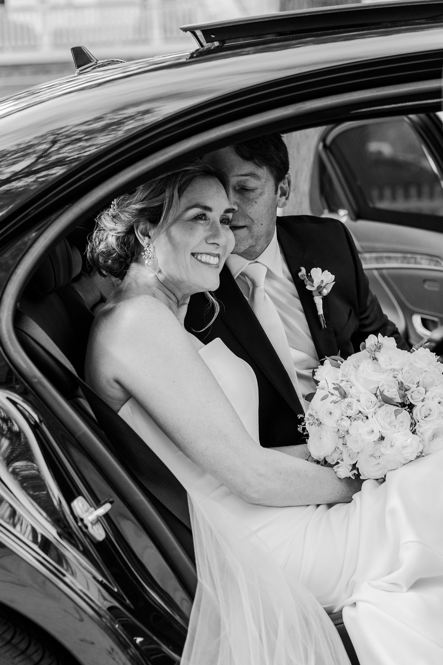 A tender black and white photograph of newlyweds sharing an intimate moment in the backseat of a car.