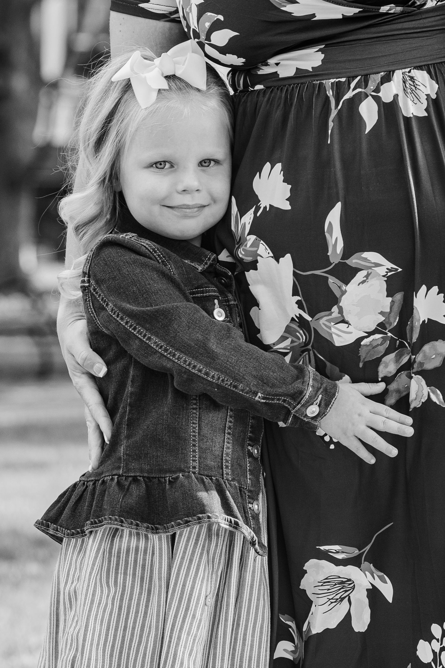 Black and white portrait of young girl in denim jacket hugging against floral dress.