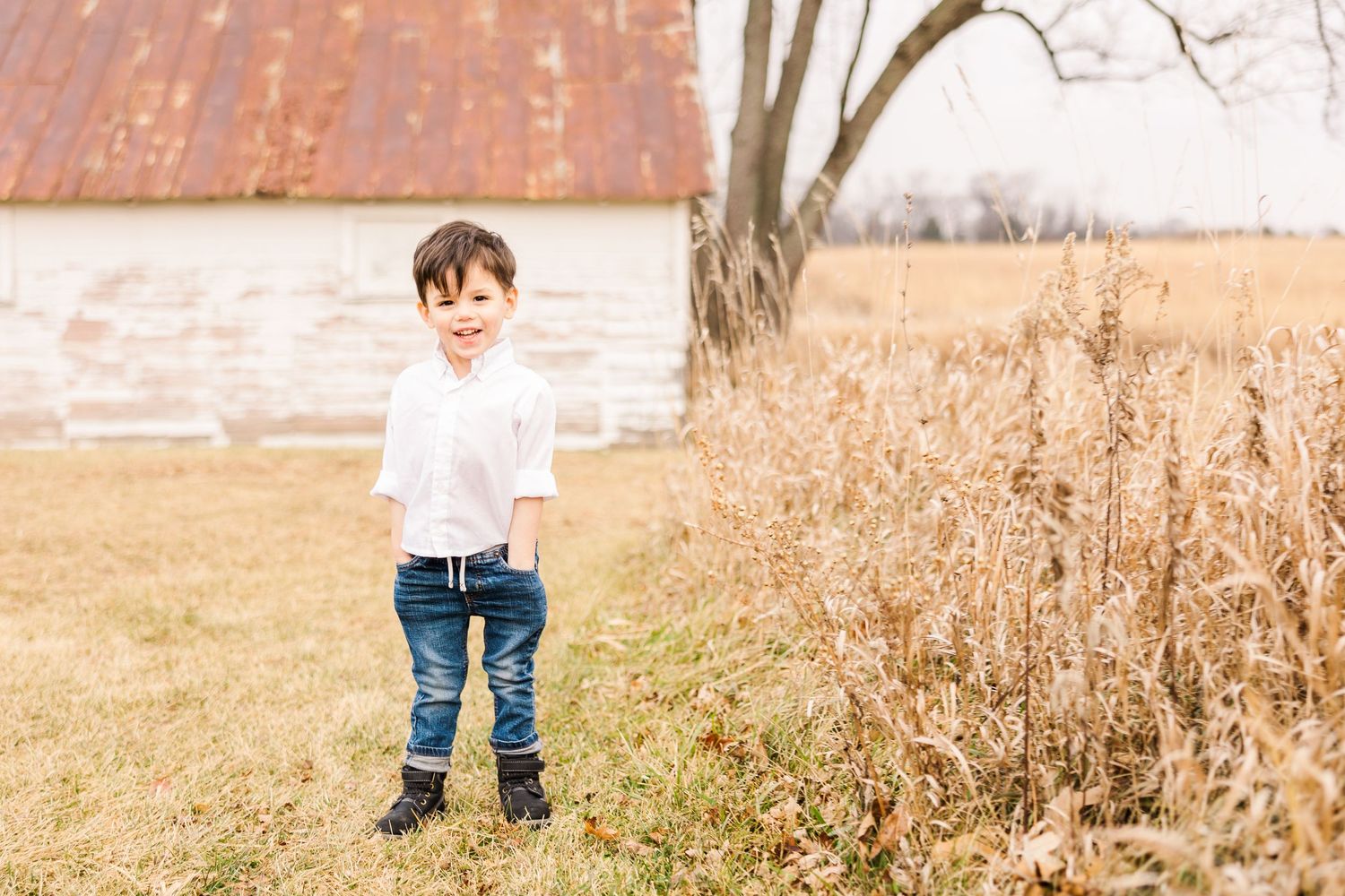 A small child stands in casual wear near a rustic barn and tall grass.