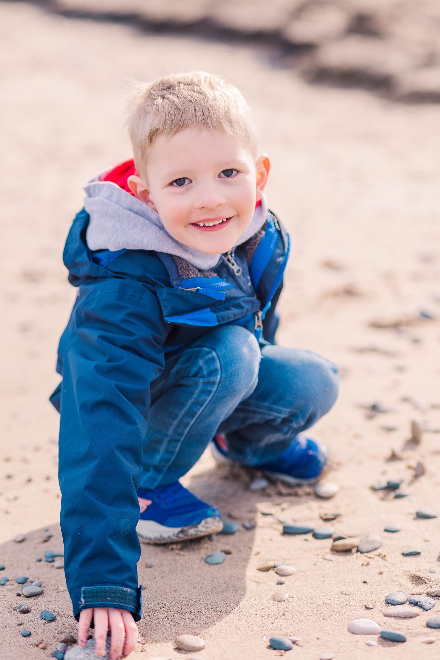 Child in blue jacket squats down to examine stones scattered on beach.