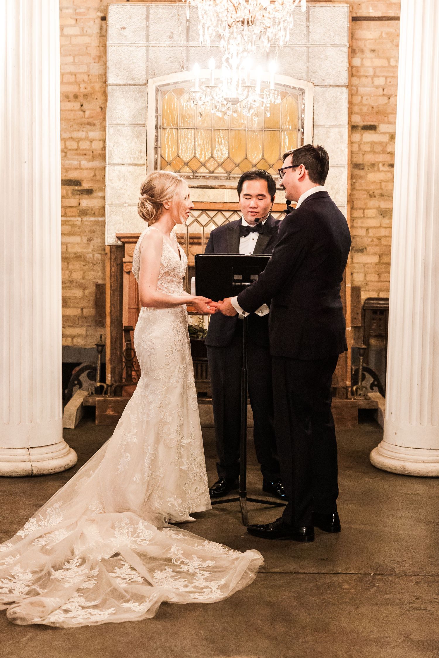 Wedding ceremony under ornate chandelier with white columns and exposed brick walls.