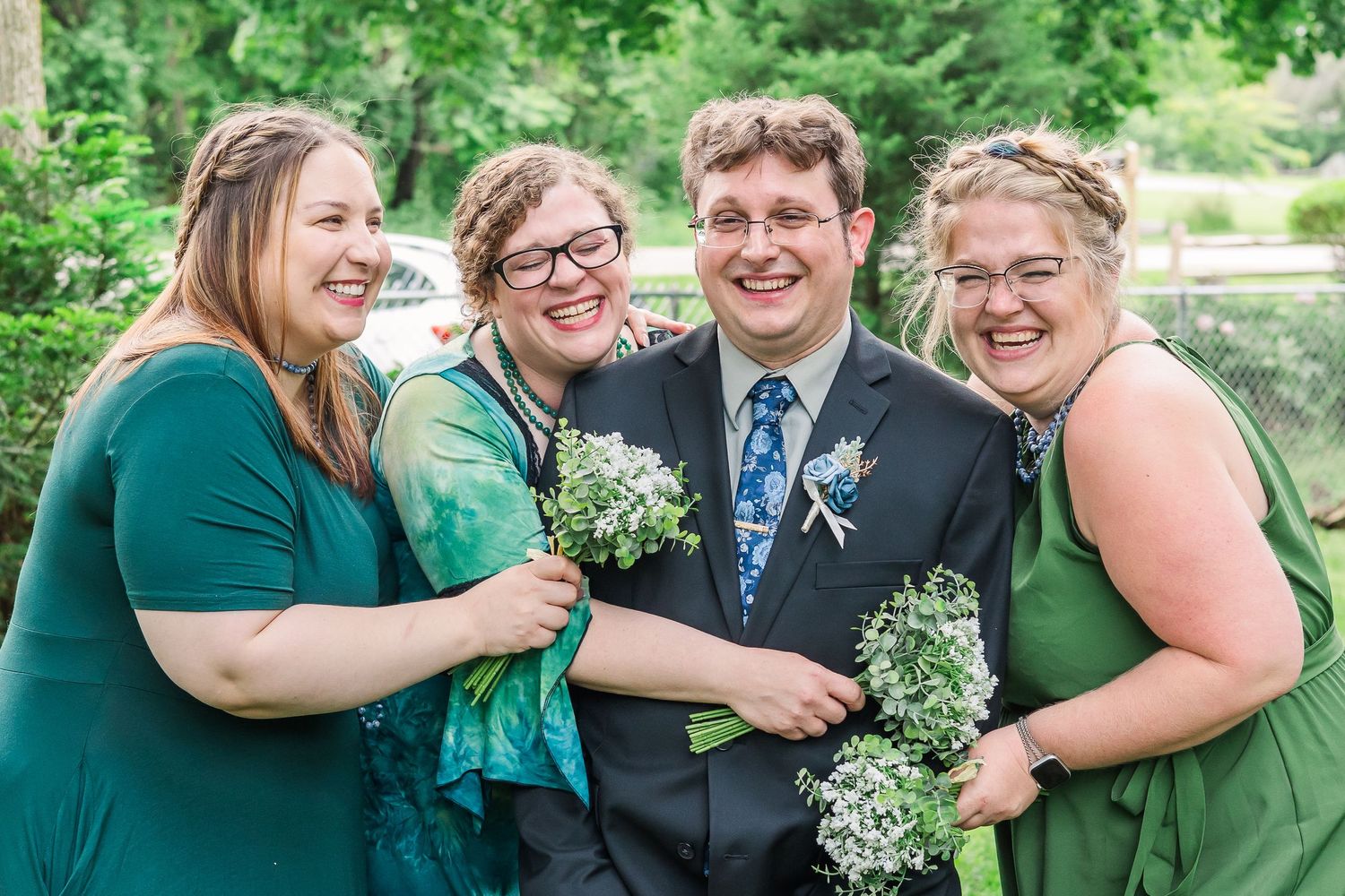 Wedding party members in green dresses and suits laugh together while holding bouquets outdoors.