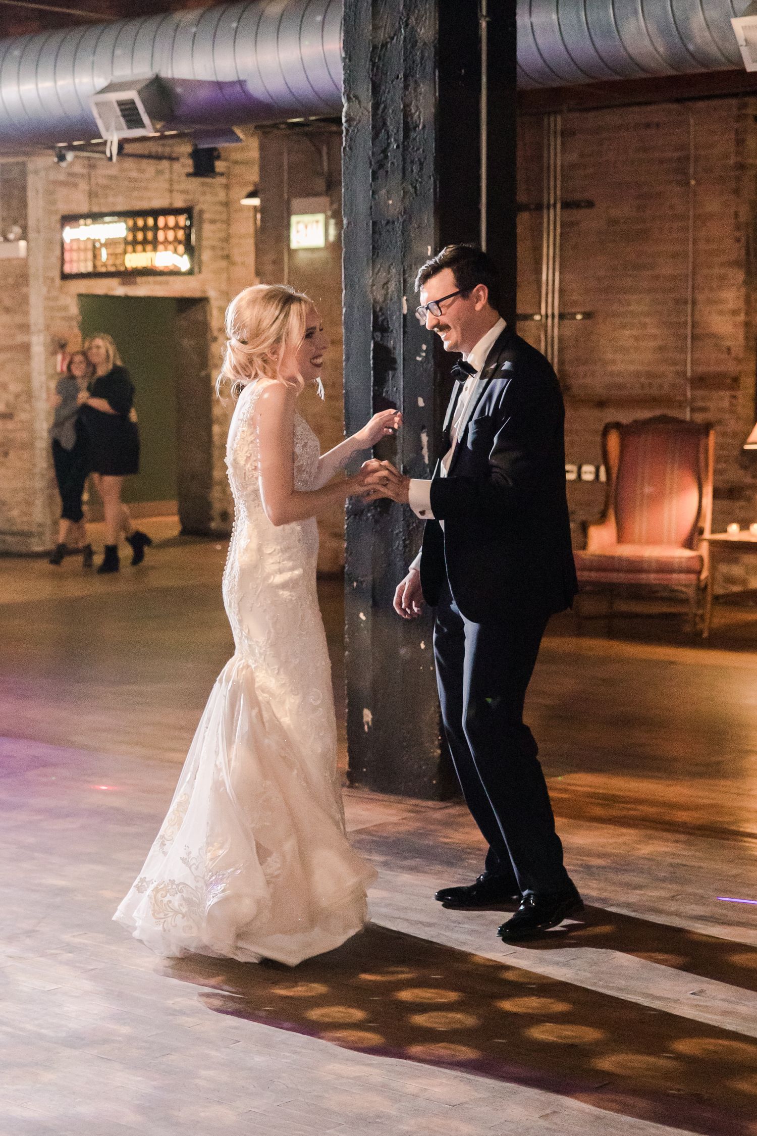 A joyful wedding dance moment in a rustic brick-walled venue with purple lighting.