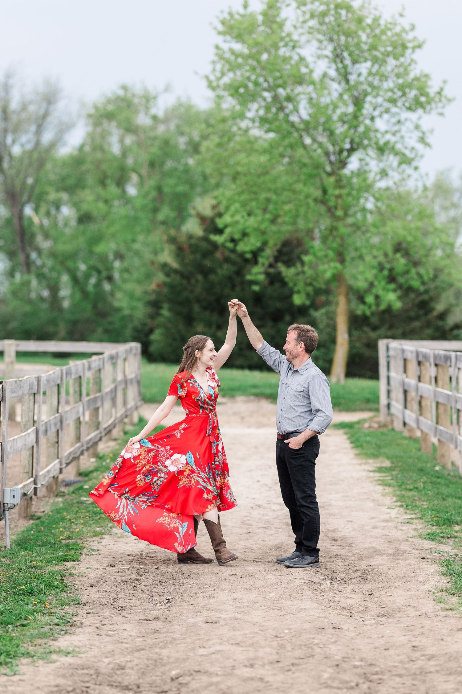 Couple dancing on a dirt path between wooden fences with green trees in background.