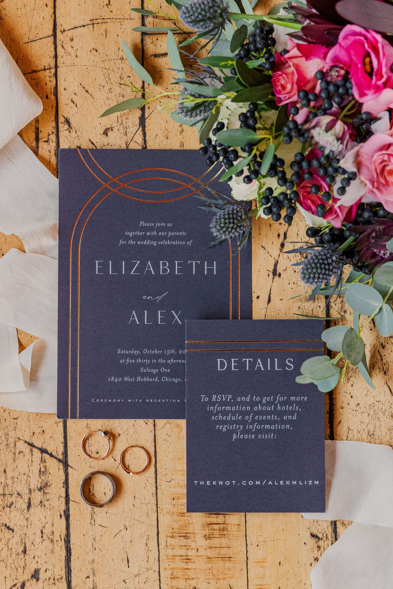 Navy blue wedding invitations with copper details displayed alongside pink roses and wedding rings.