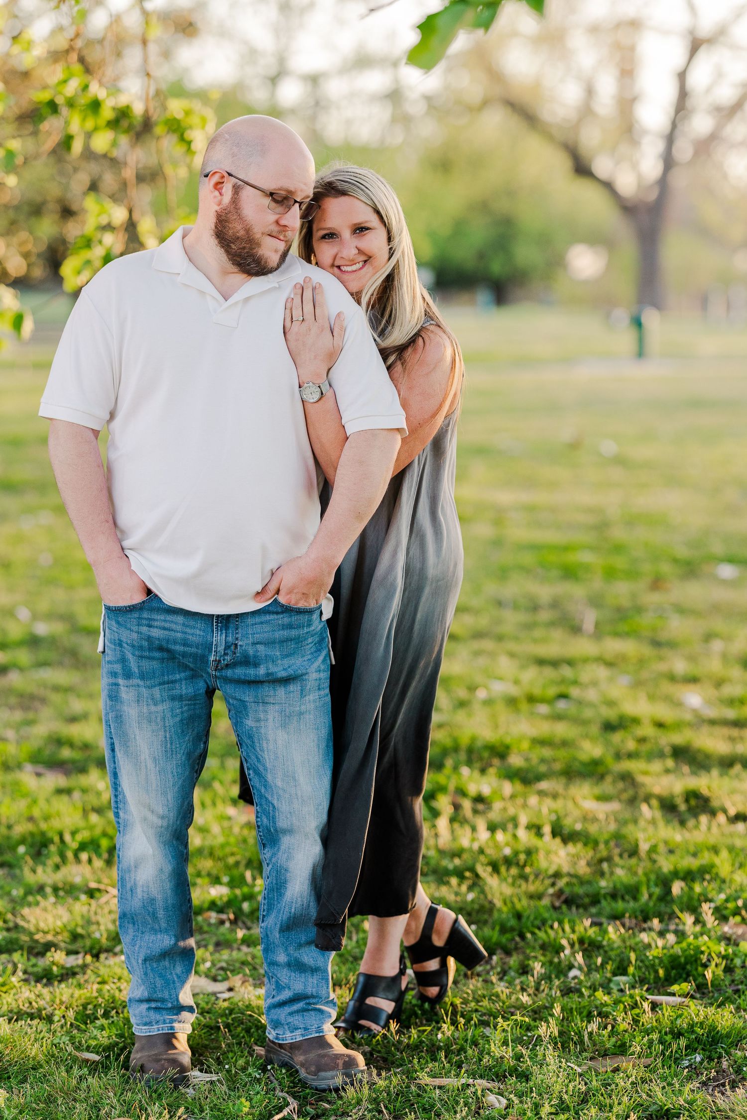 A couple poses playfully in a park with green trees and grass in the background.