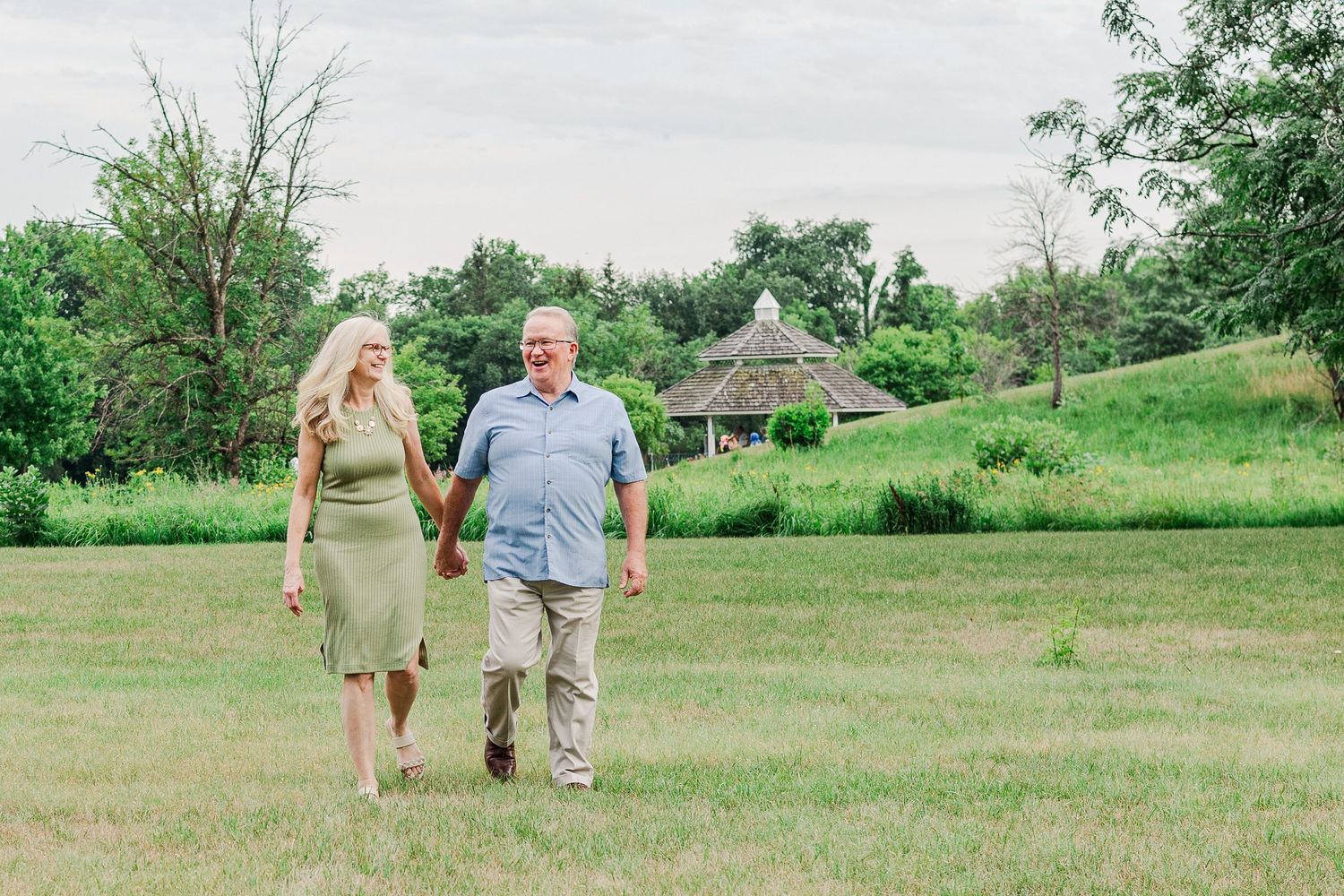 A couple walks hand in hand across a lush green lawn on a summer day with trees and buildings in the background.