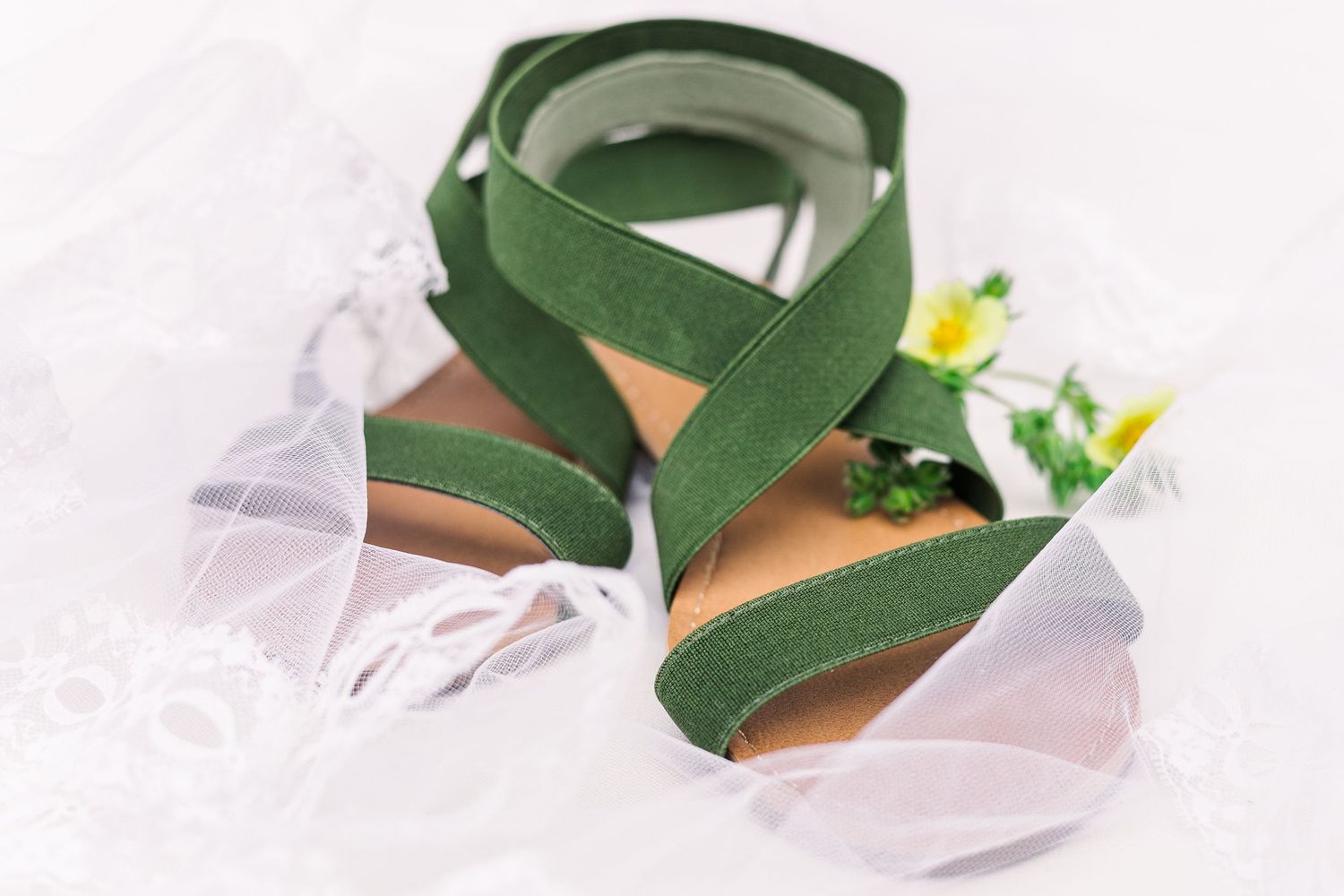 Green strappy sandals with floral accents lying on white tulle fabric.