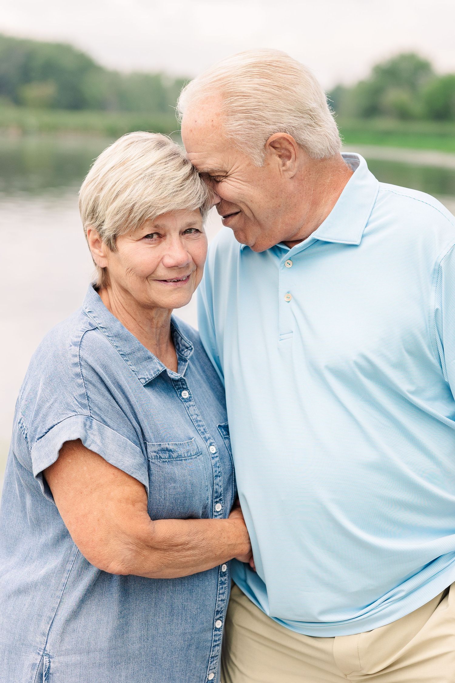 An affectionate couple embraces while standing by a lake, wearing casual blue and khaki clothing.