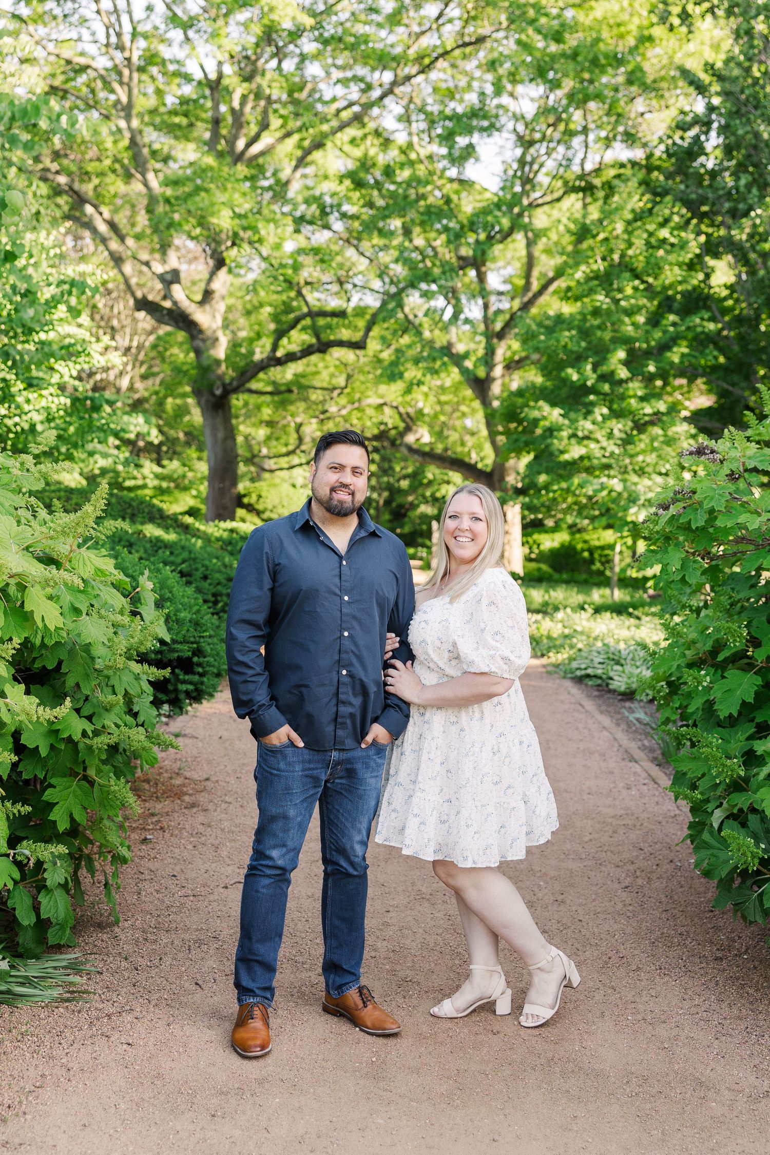 A couple poses on a garden path surrounded by vibrant green foliage and trees.