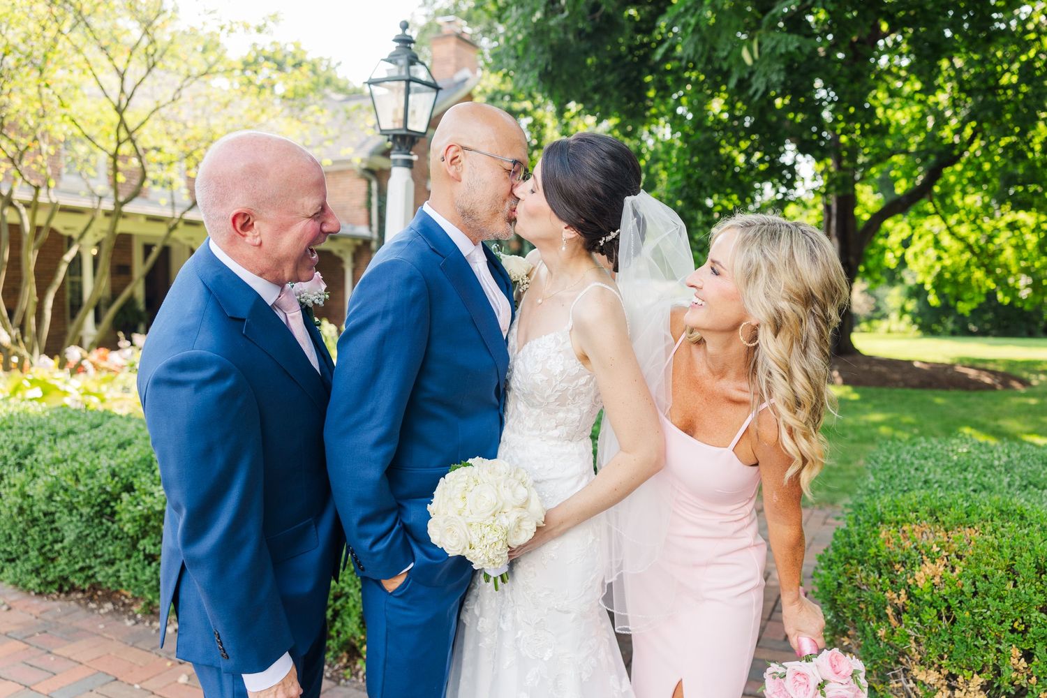 Wedding party in blue suits and pink dresses gathered outdoors in a garden setting.