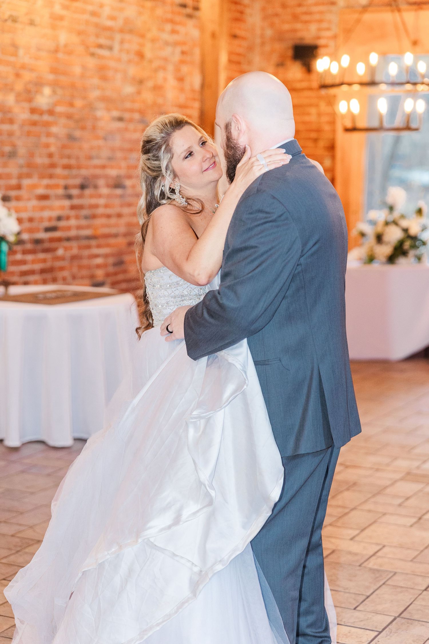 A bride and groom share their first dance in a rustic brick venue with elegant lighting and floral decor.