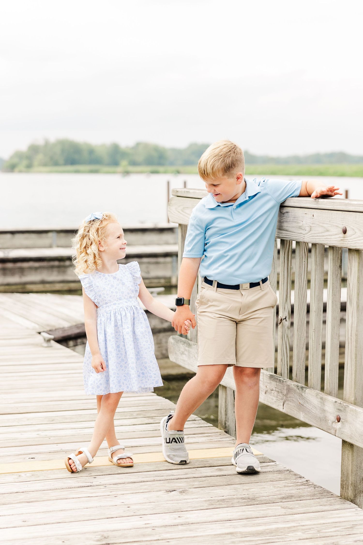 Two children holding hands while standing on a wooden lakeside dock on a bright summer day.