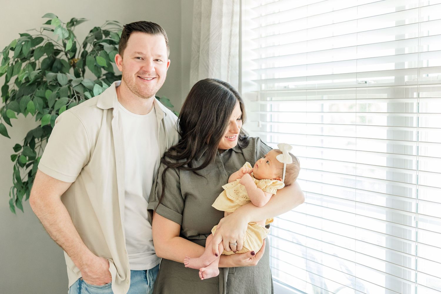 Dad smiling as mom snuggles baby girl by the window during a lifestyle newborn session at home