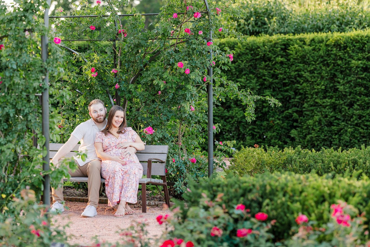 Wide garden portrait of expecting parents seated beneath a rose arch at Cantigny Park in Wheaton Illinois