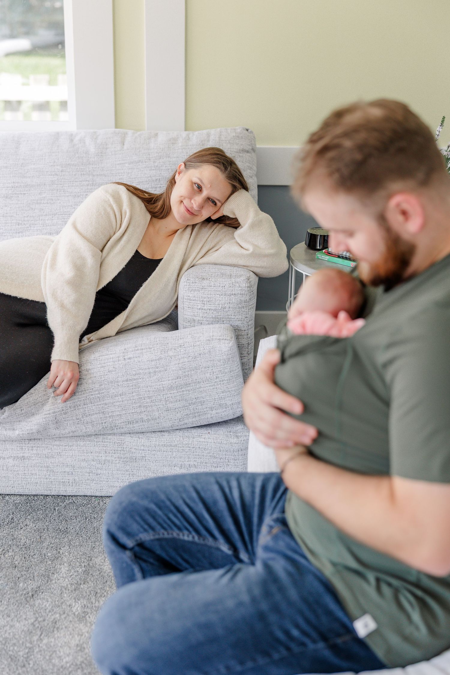 A mother relaxes on a gray sofa smiling as a bearded father sits on the floor cradling their newborn baby.
