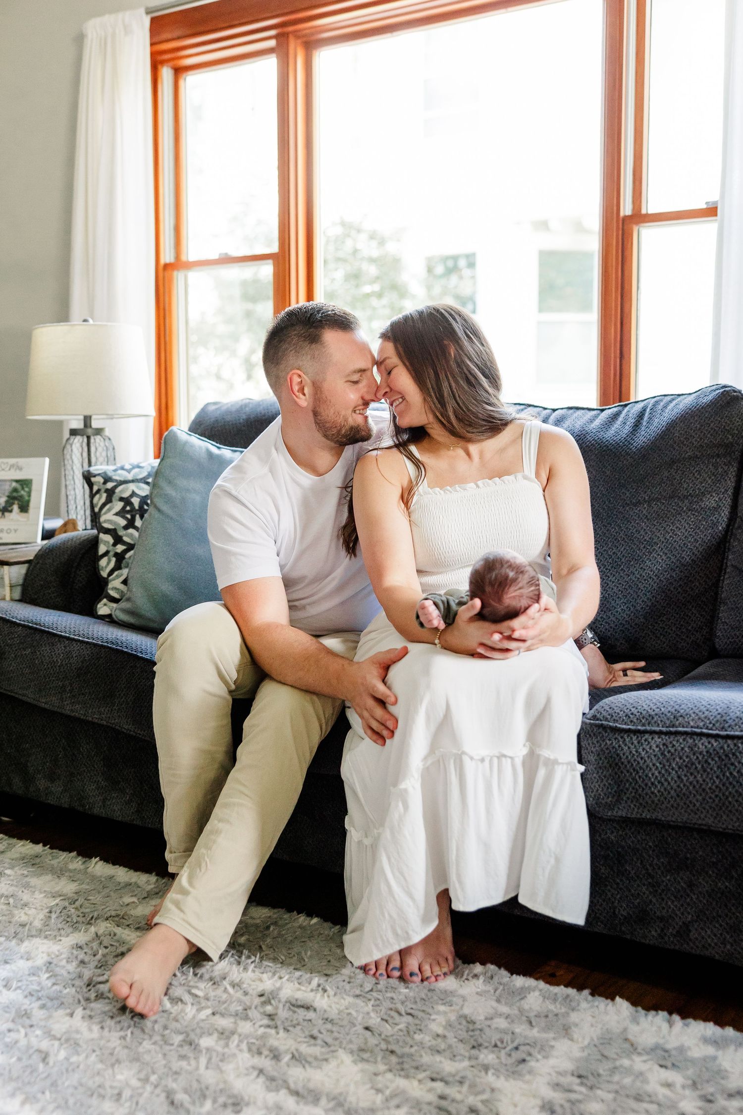 Happy couple sharing a tender moment on a gray sofa while cradling their newborn baby in a cozy living room.