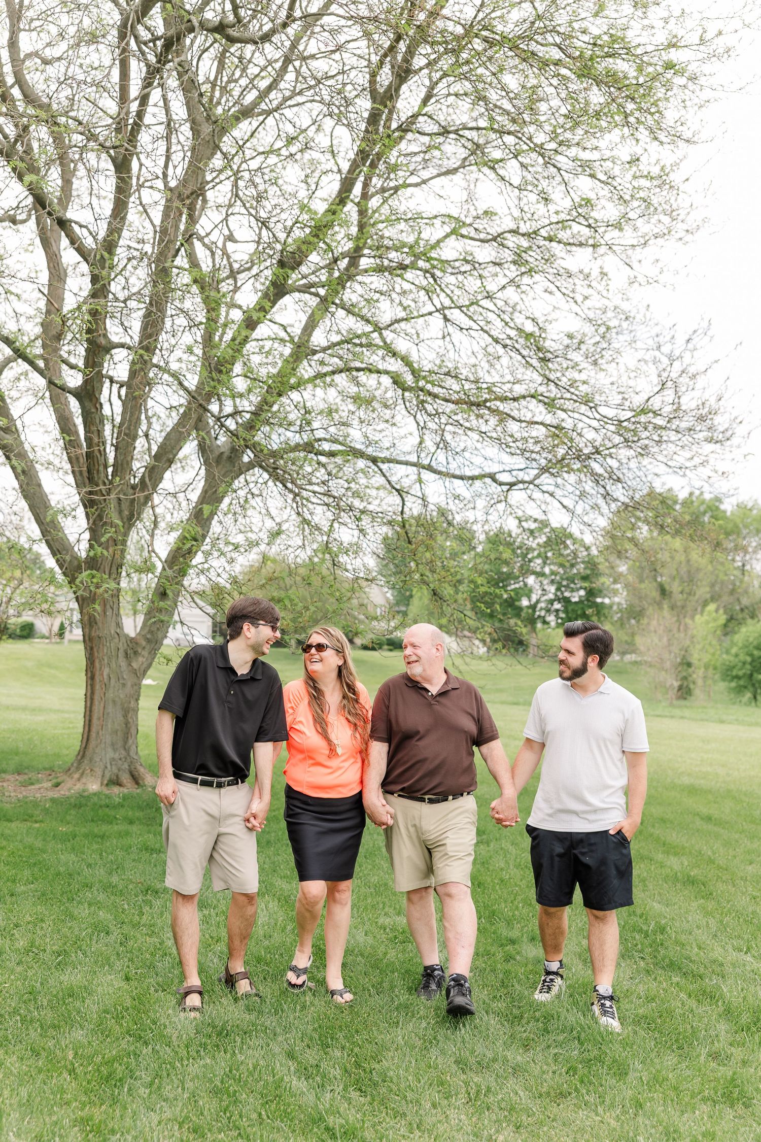 Family of four walks hand-in-hand beneath a large tree on green grass during an outdoor portrait session.