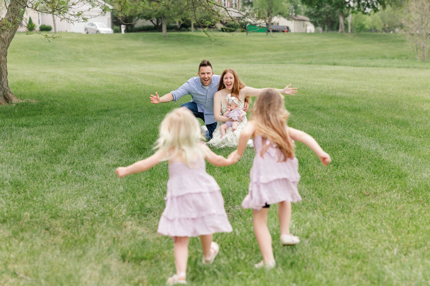 Two young girls in pink dresses run toward their parents with open arms on a lush green lawn.