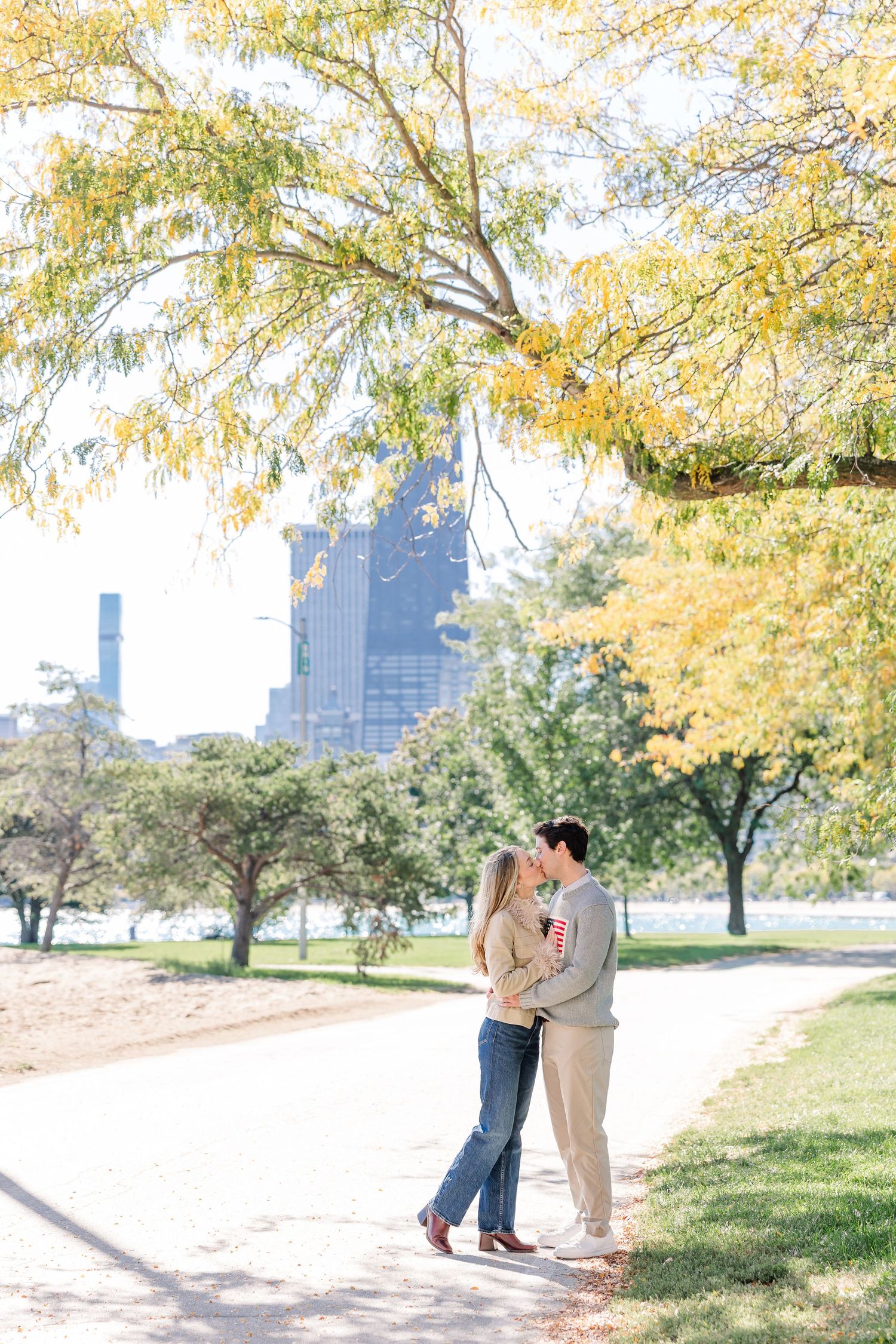 Couple embracing on a park path surrounded by autumn yellow trees with a city skyline in the background.
