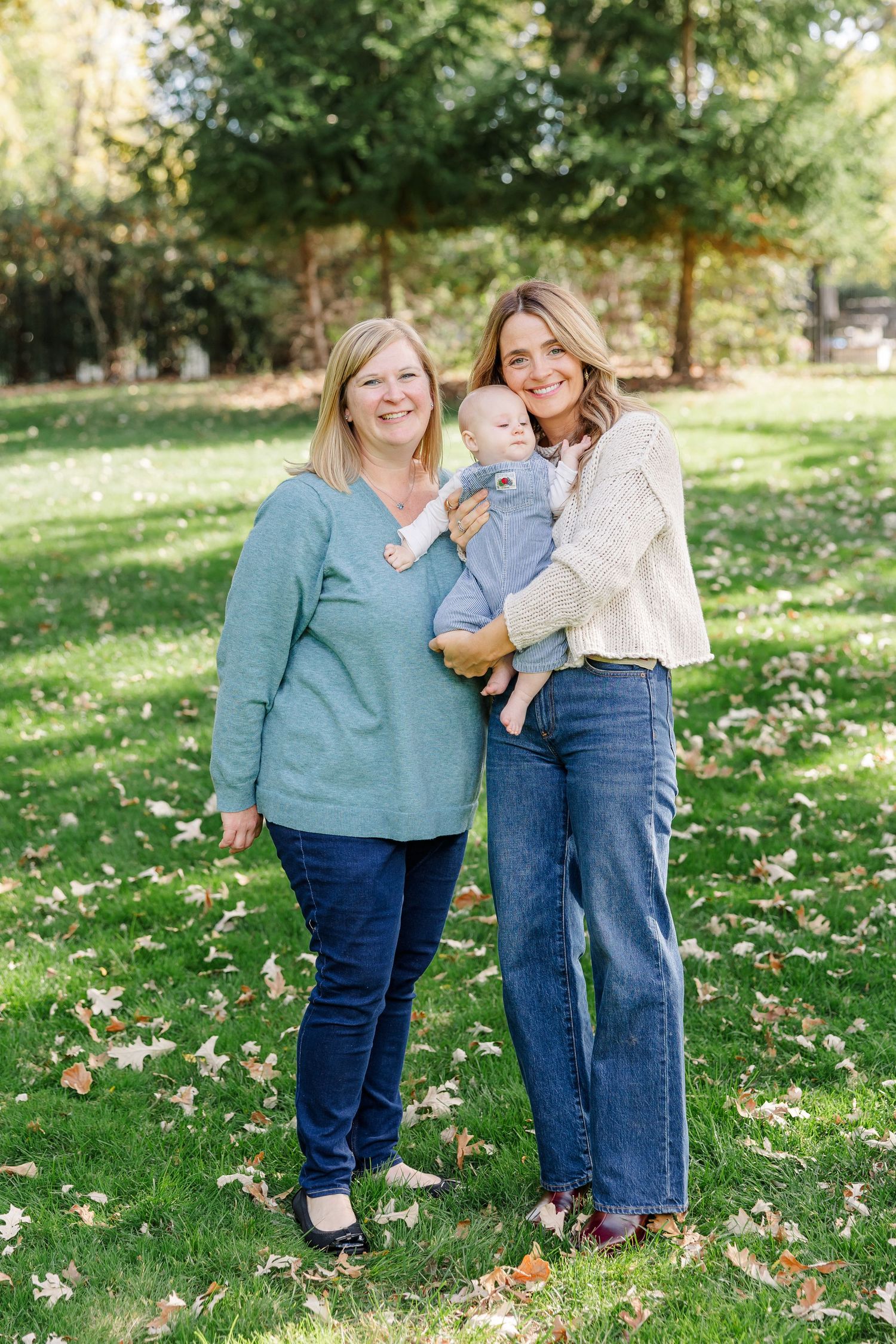 Two women and a baby posing outdoors on green grass with autumn leaves, surrounded by trees in a park setting.
