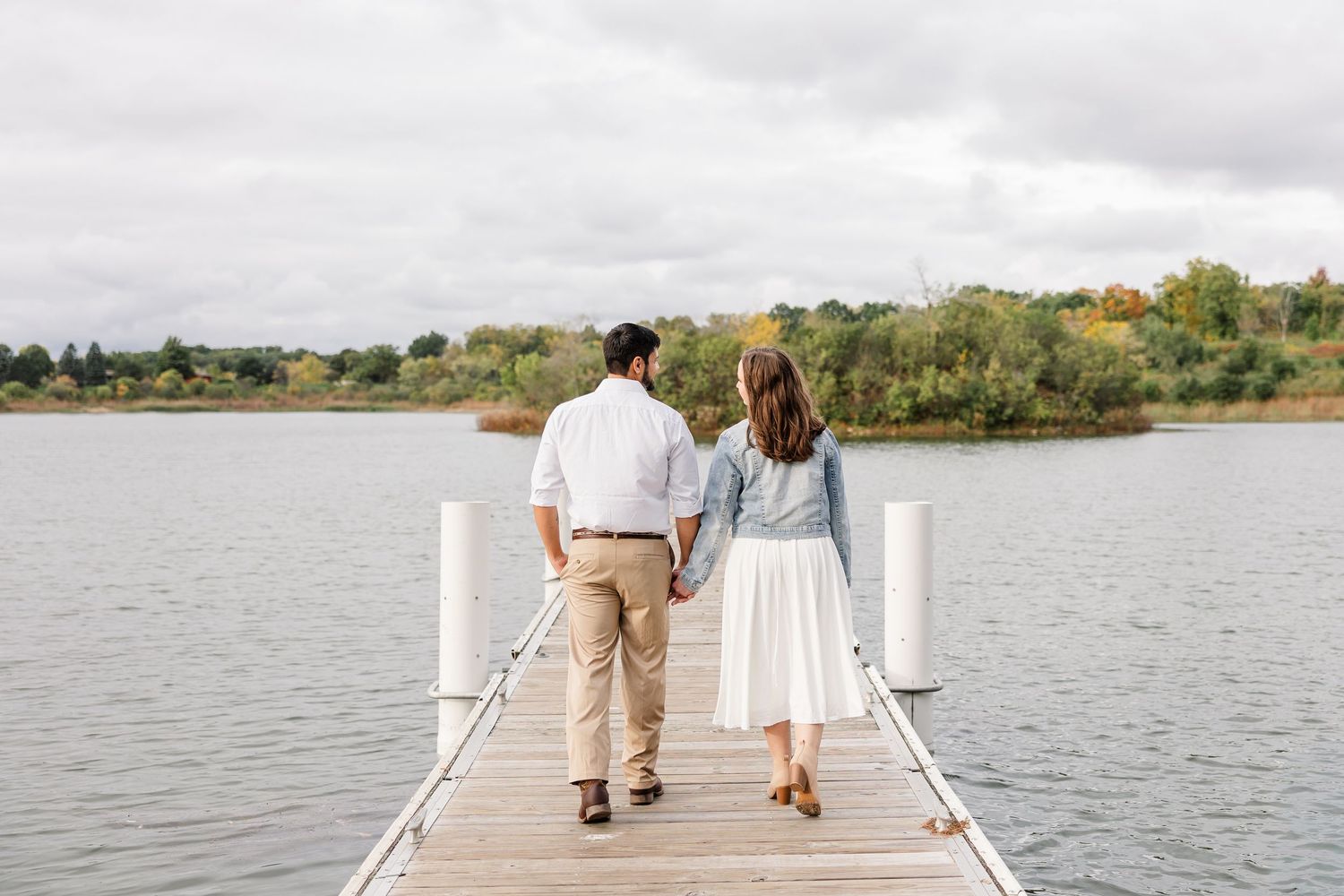 Couple holding hands walking away on a wooden dock over a calm lake with autumn trees and a cloudy sky in the distance.