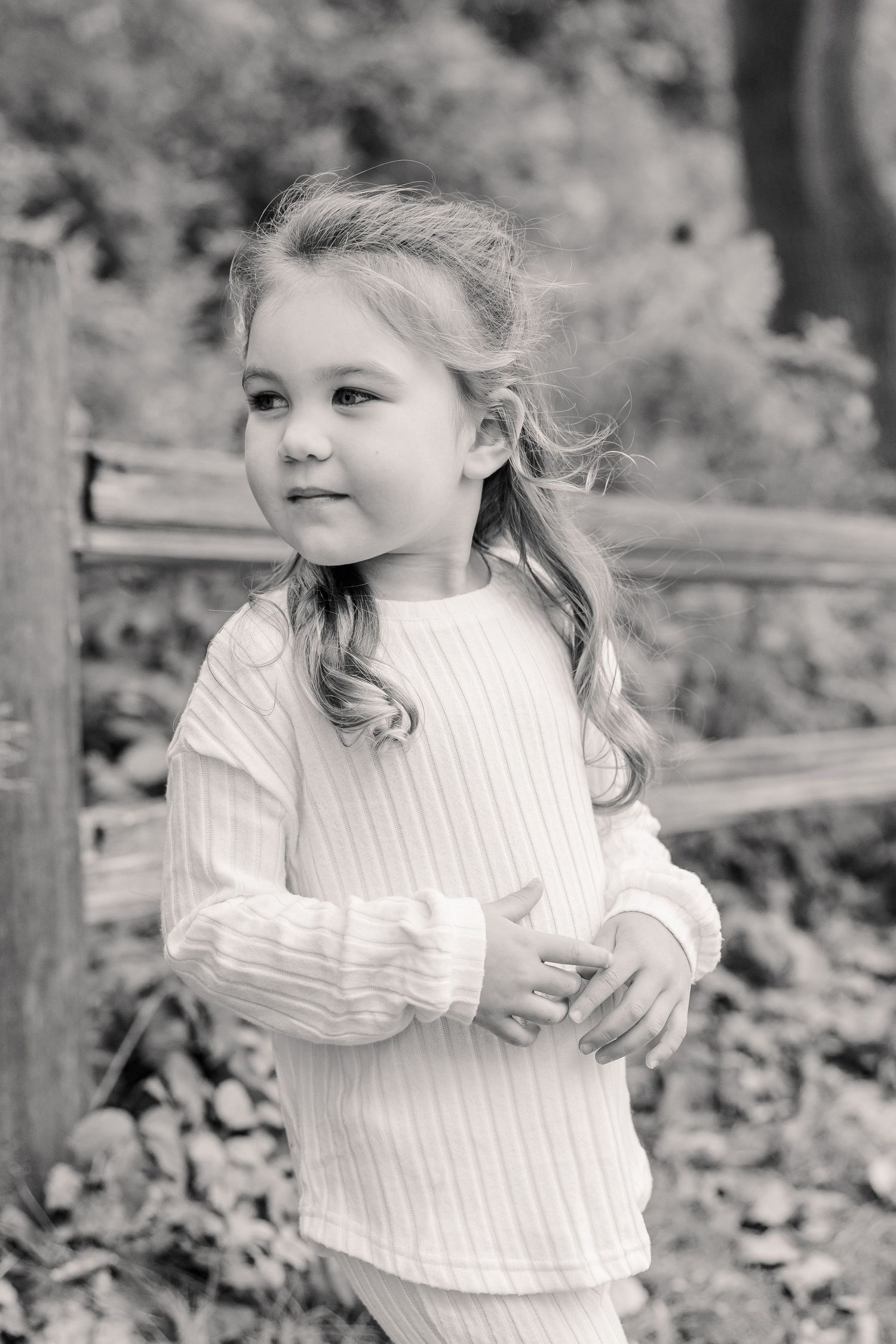 A sweet young girl with long hair poses outdoors in a white outfit in this charming black and white portrait.