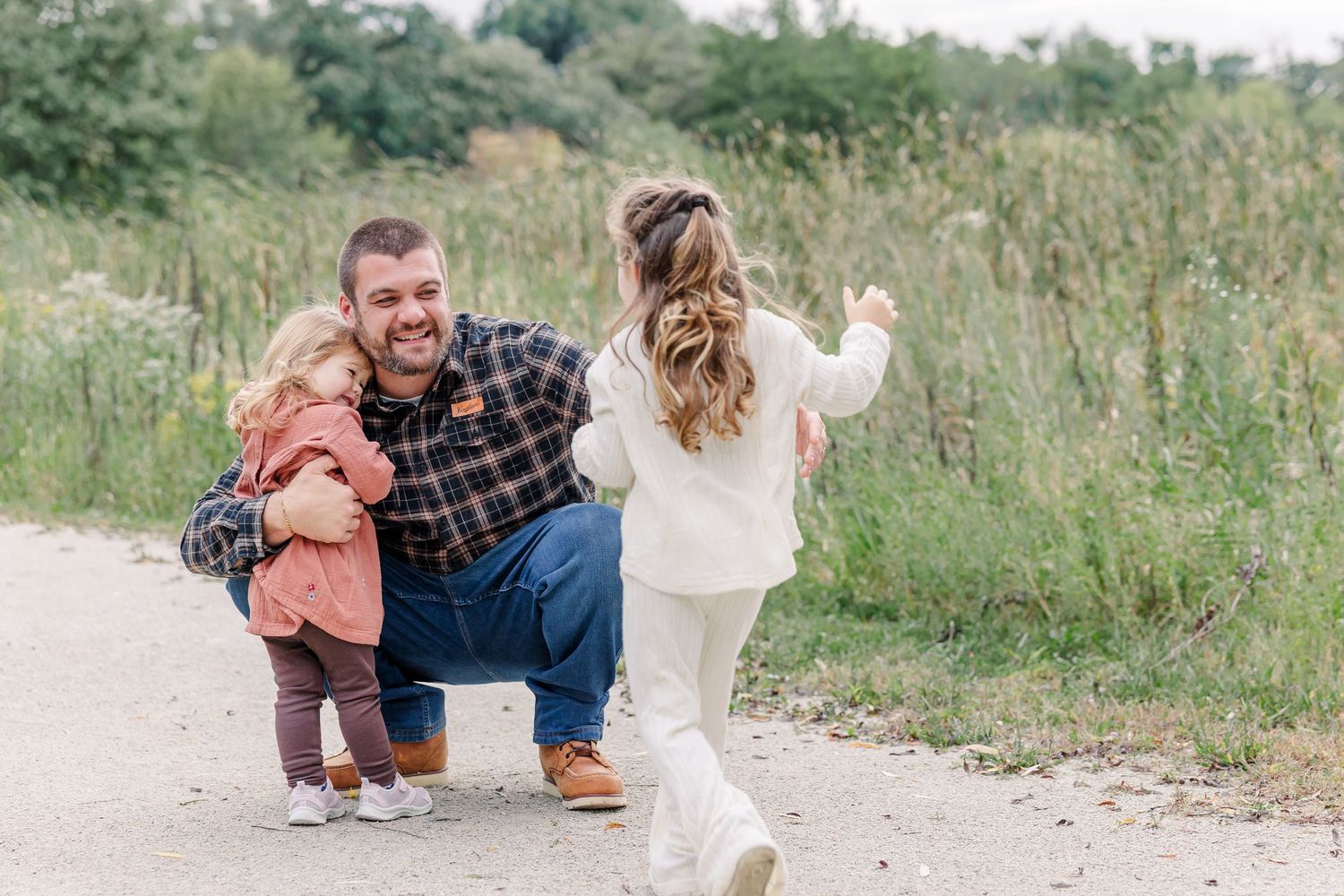 Father crouching on a path hugging a toddler while a young girl runs toward them outdoors.