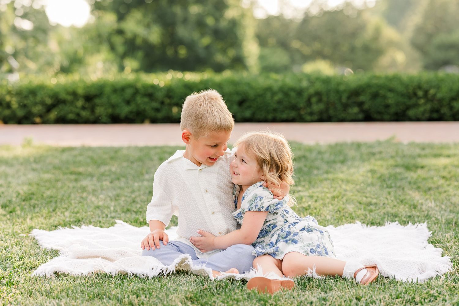 Two young children sit together on a white blanket in a lush green park, sharing a sweet moment.