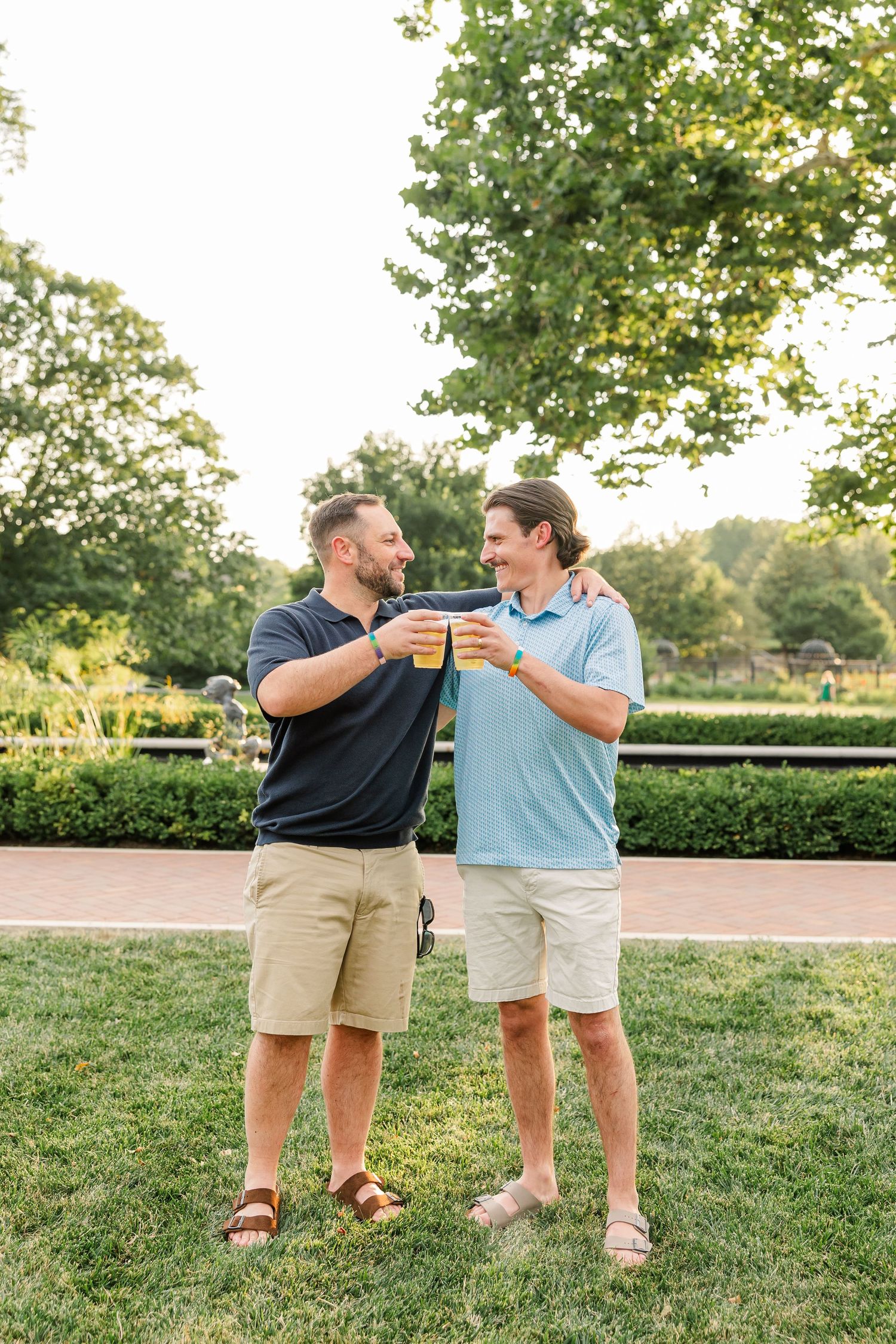 Two men toasting with orange drinks on a green lawn in a sunny park, smiling at each other in casual summer outfits.