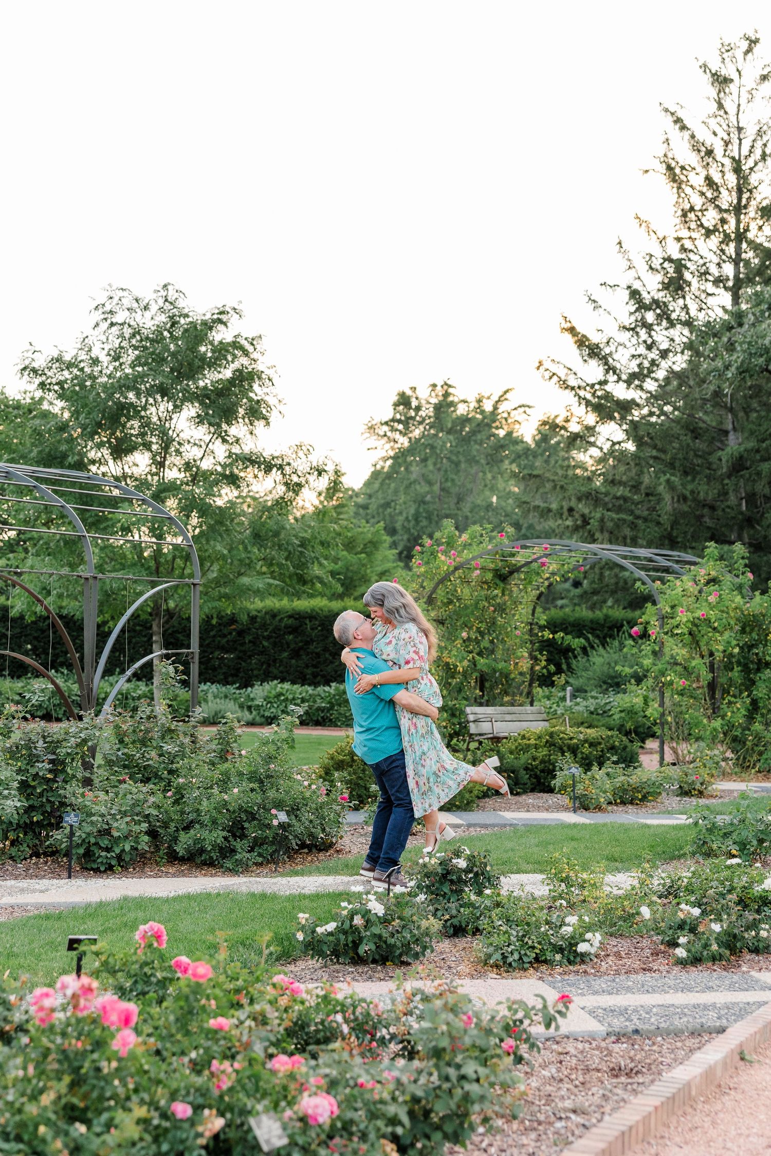 Couple sharing a romantic lift and embrace among blooming roses in a lush garden at golden hour.