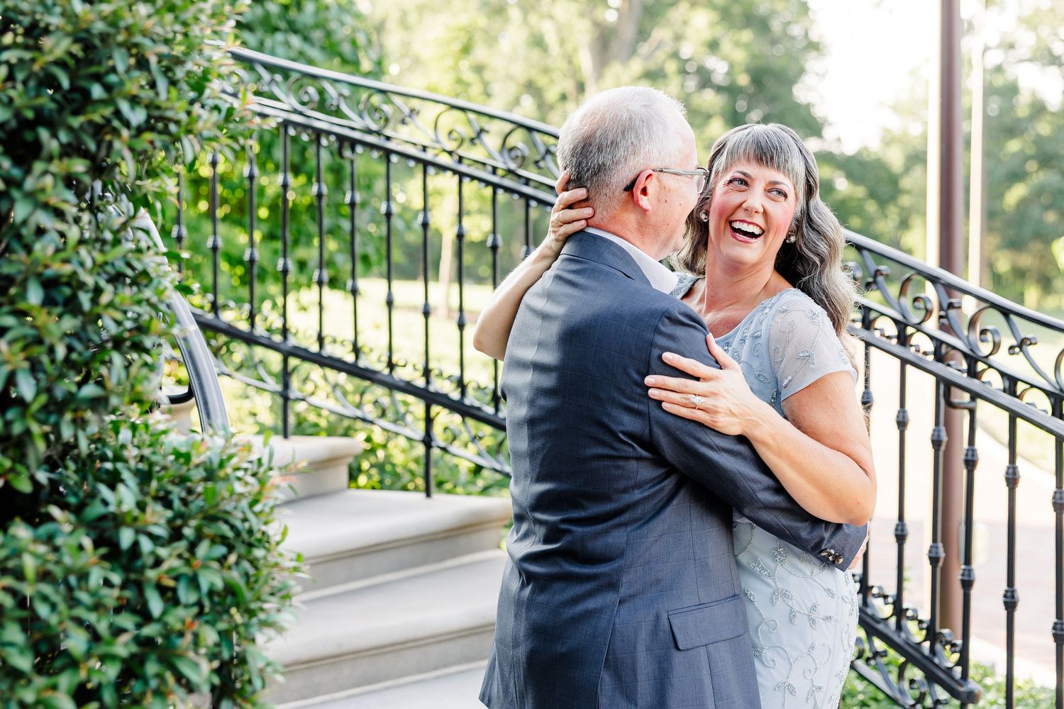 Older couple embracing and laughing together on outdoor stone steps with black iron railing and green foliage.
