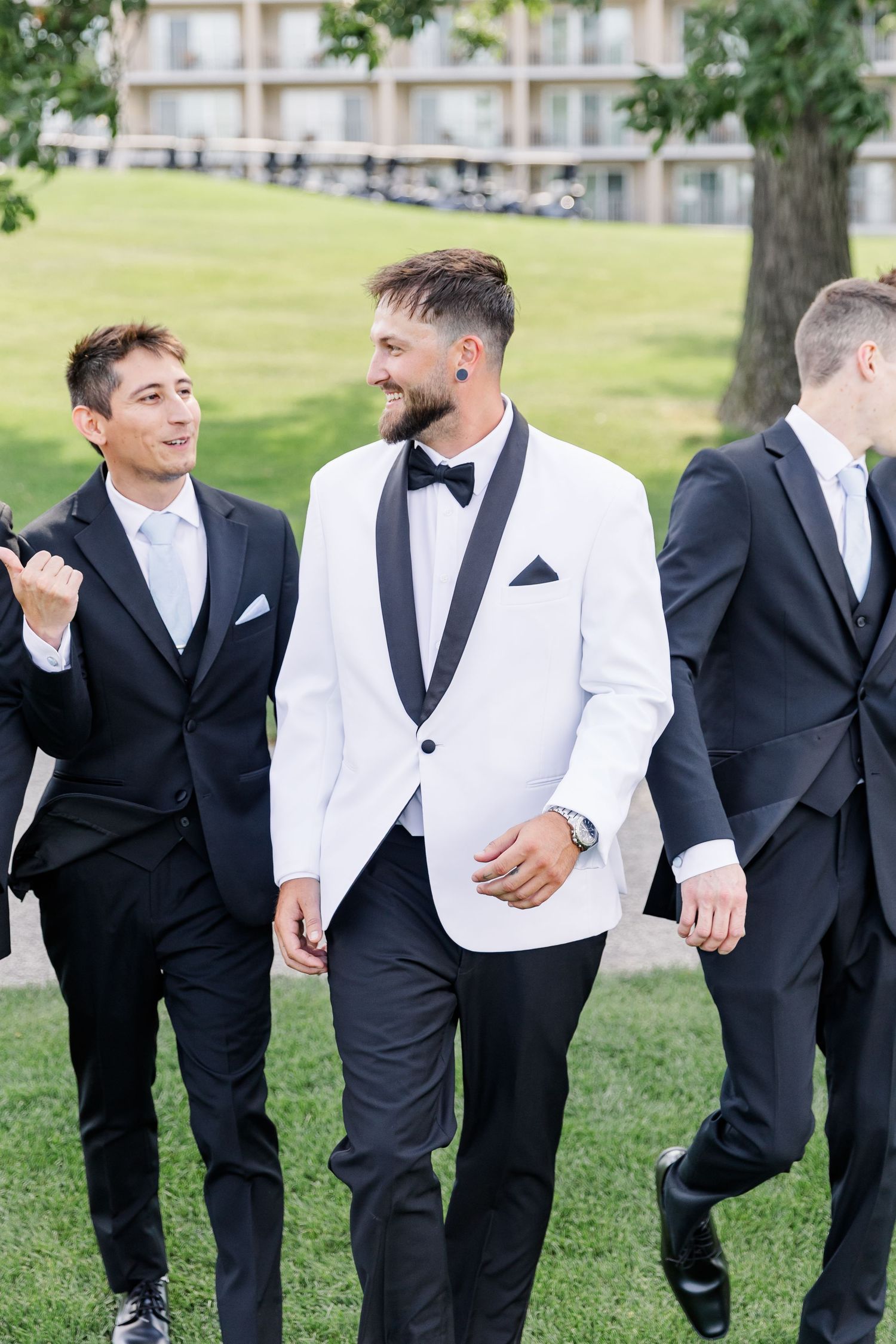 Groom in white tuxedo jacket walks and laughs with groomsmen in black suits outdoors.