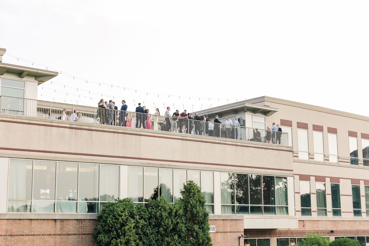 Wedding guests gathered on rooftop terrace of modern building with string lights above.