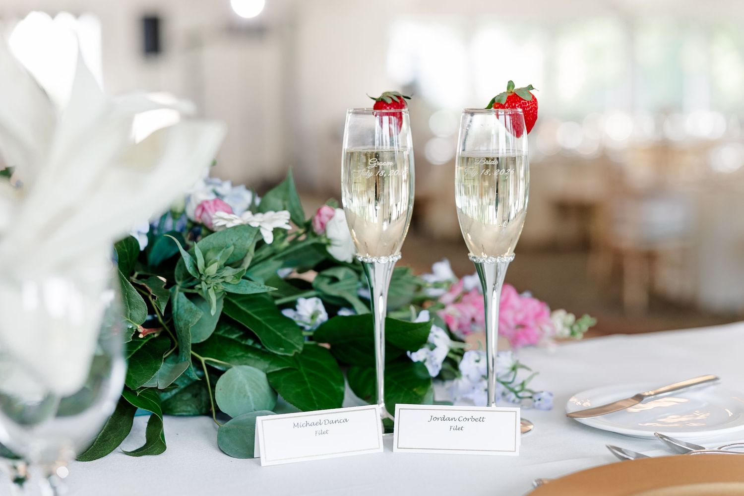 Two champagne flutes garnished with strawberries sit beside a floral centerpiece on a white wedding table.