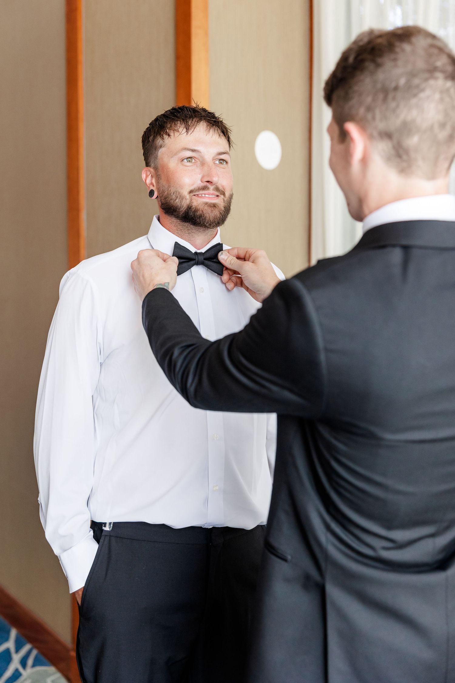 Groomsman adjusting groom's black bow tie as he smiles in white dress shirt.