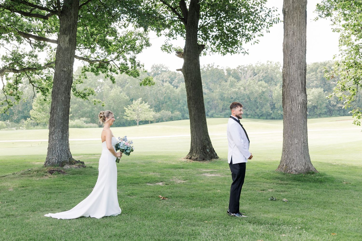 Bride and groom share an outdoor first look moment surrounded by tall trees on a green lawn.