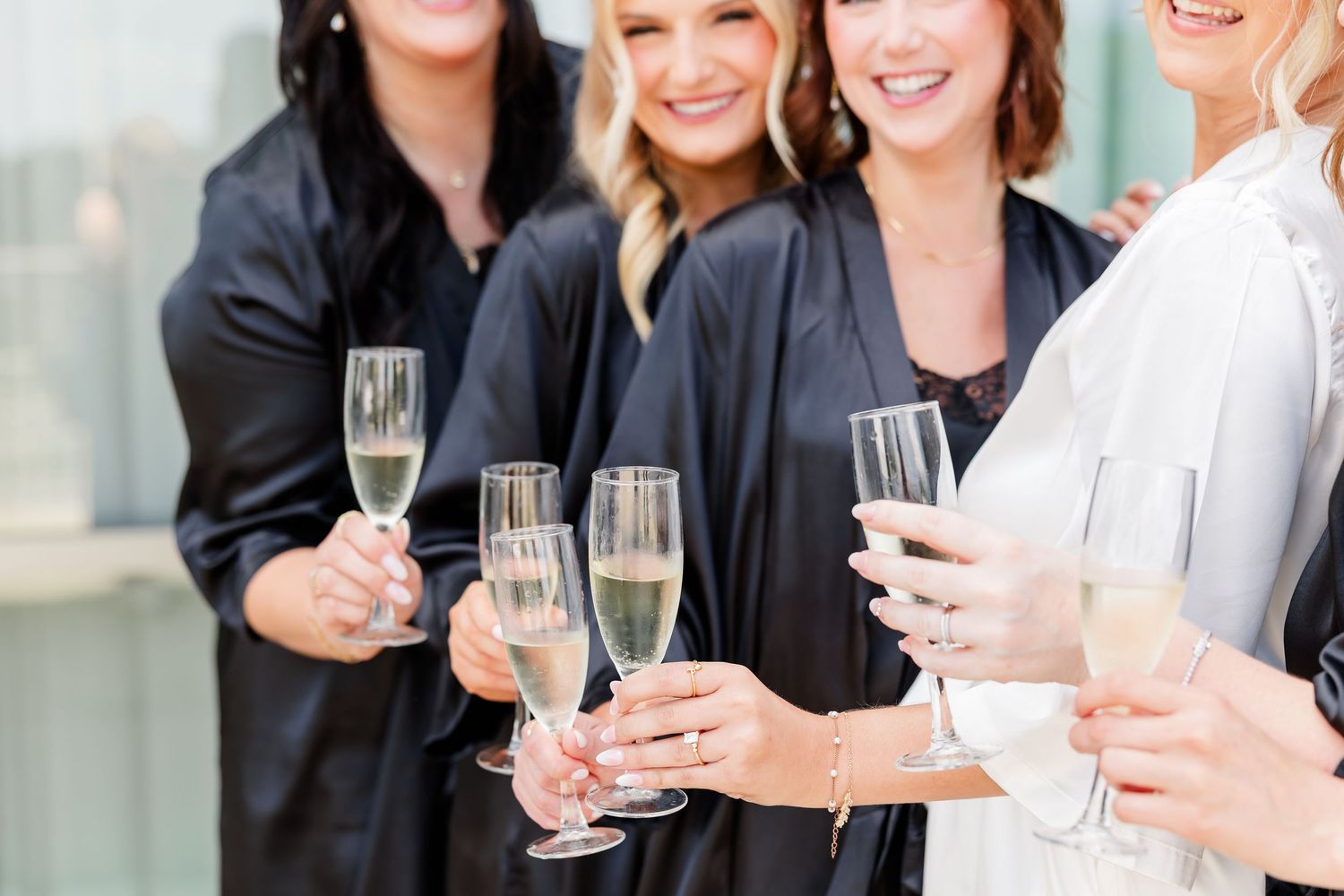 Bridesmaids in black and white robes toasting with champagne flutes during bridal party preparations.