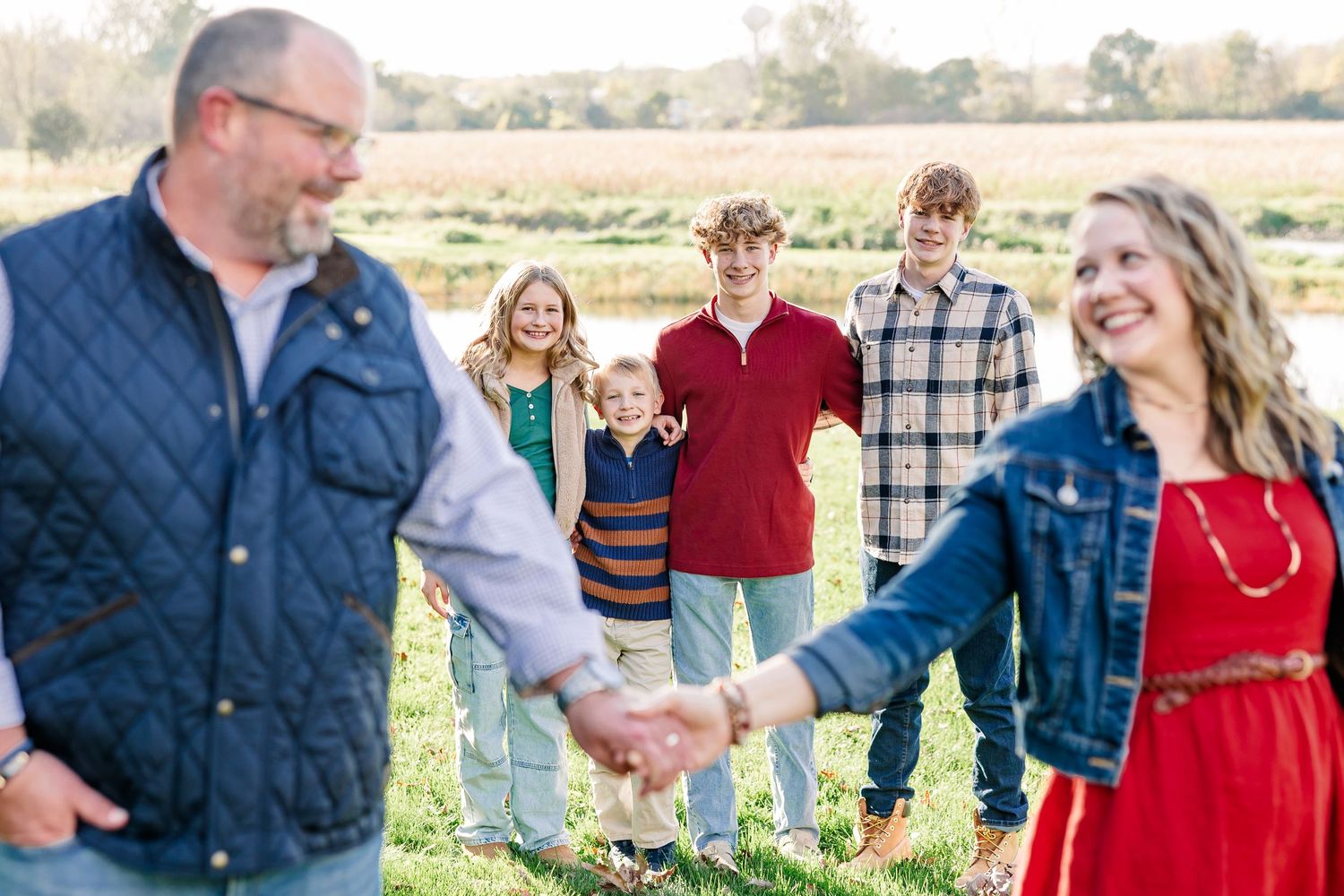 Smiling family of six holding hands in a grassy field, with a man and woman in the foreground and four children standing in the background.