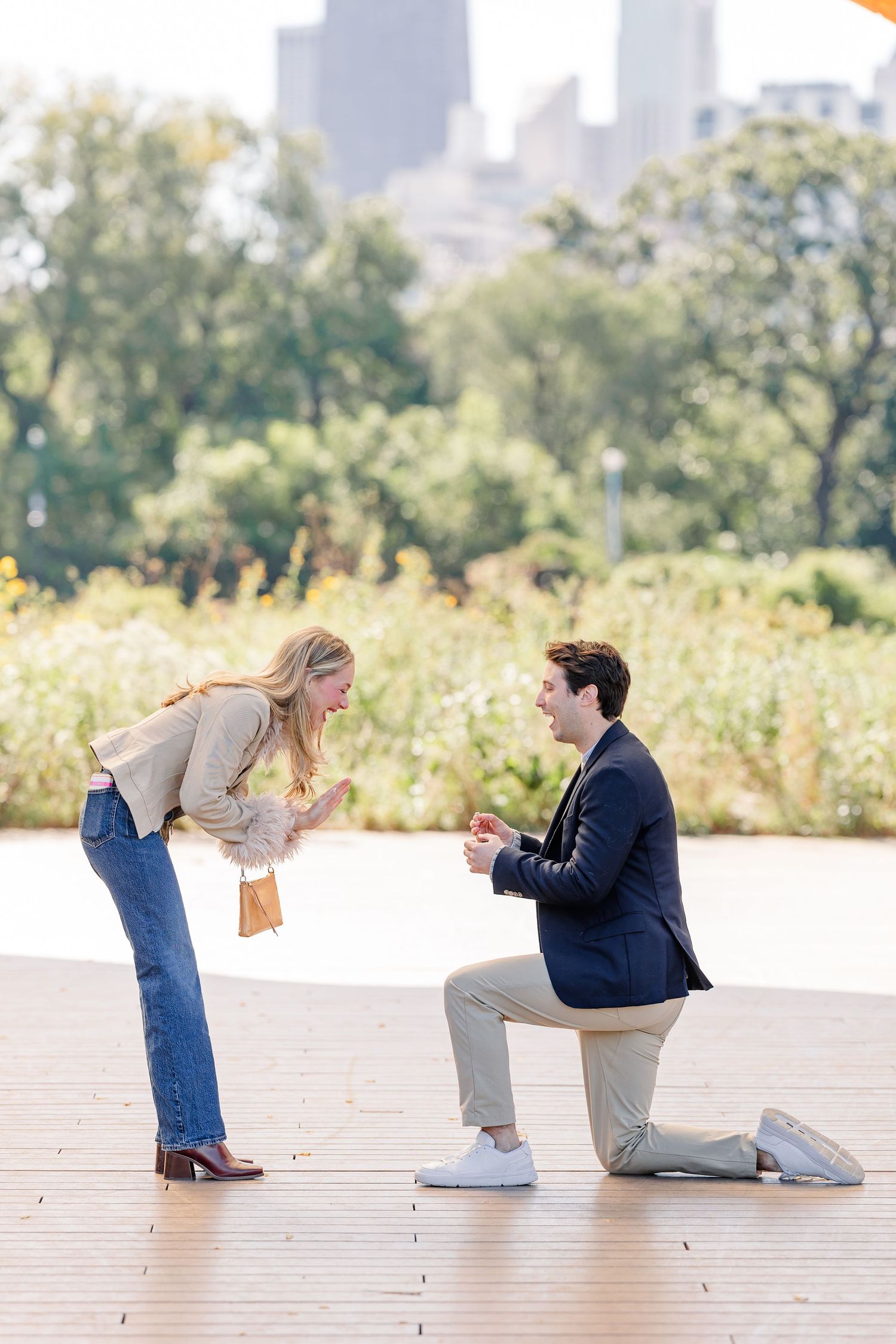 Man proposes on one knee to woman in park with city skyline backdrop.