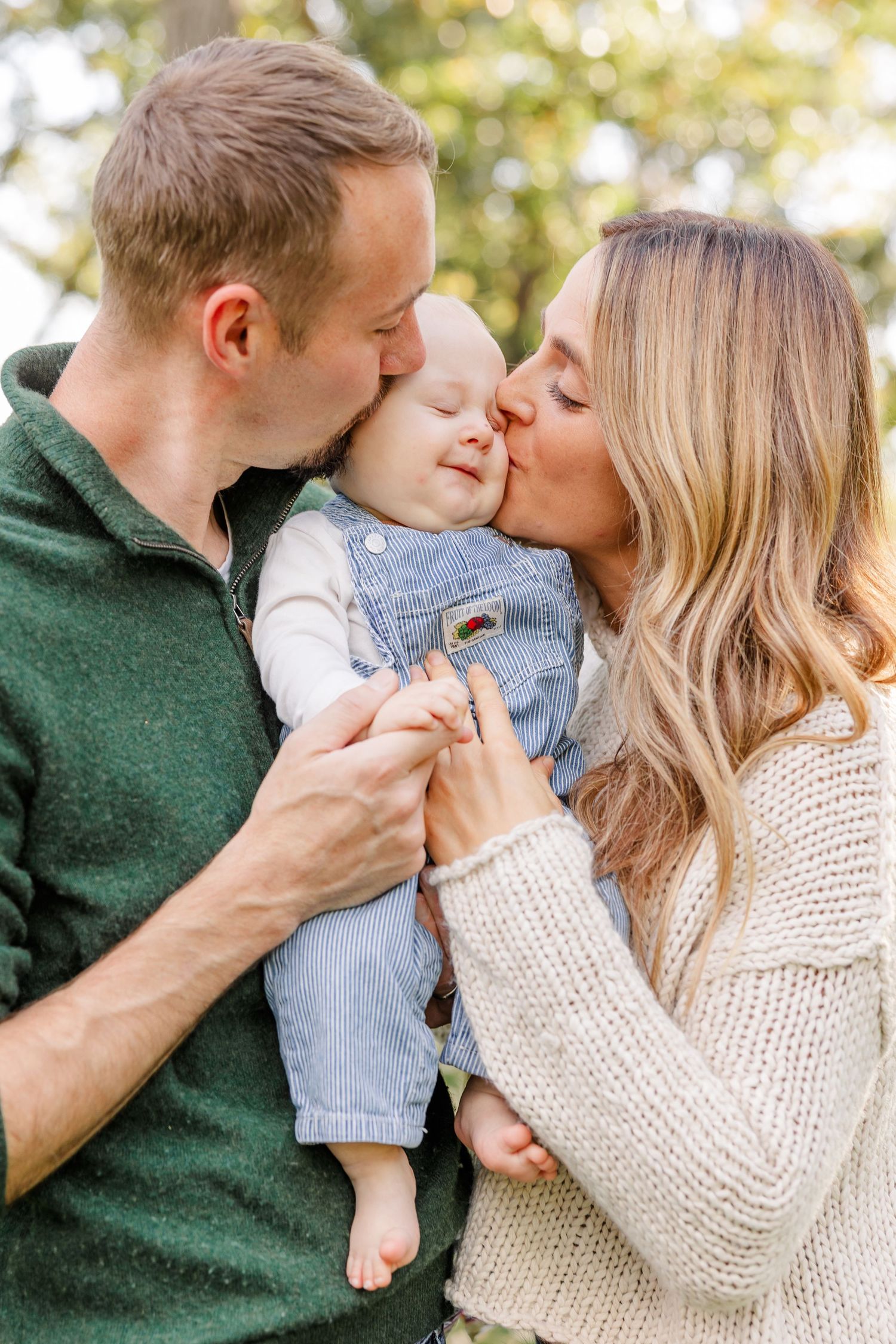 Parents kissing their smiling baby in denim overalls outdoors during a family portrait session.