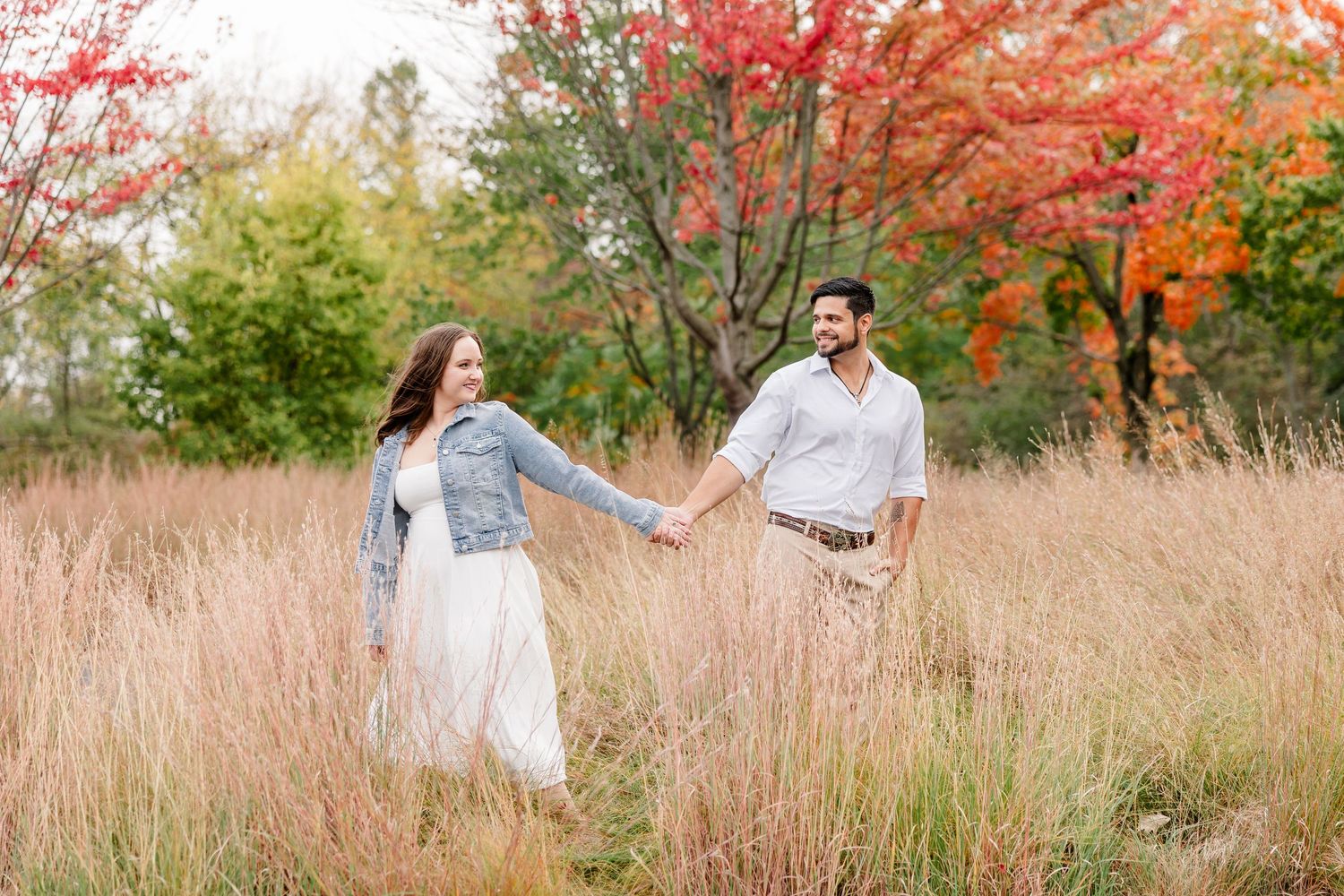 Smiling couple holding hands and walking through tall golden grass with colorful red and orange autumn trees behind them.