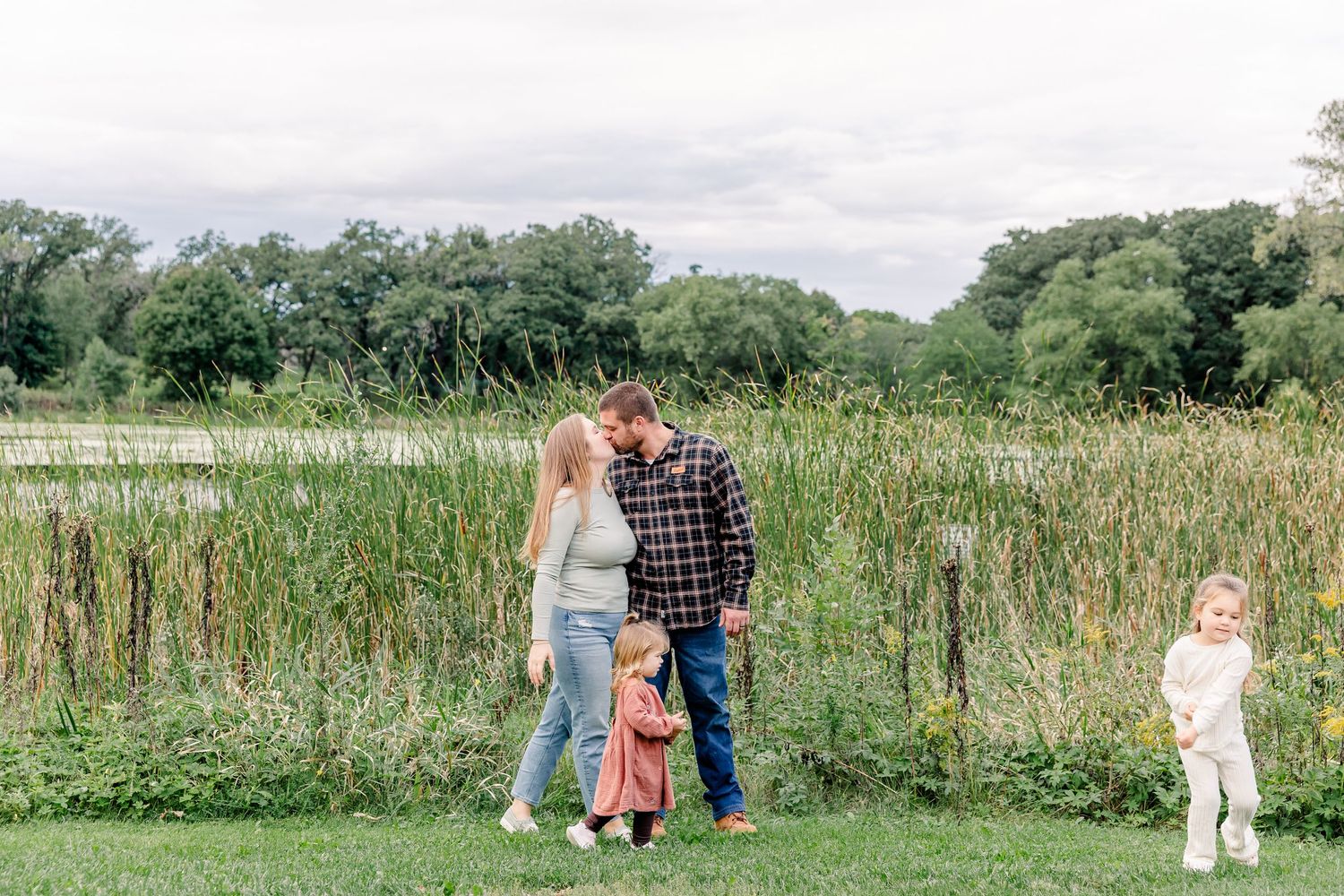 Young family of four walking together near a pond with tall reeds, sharing a tender moment in a green outdoor setting.