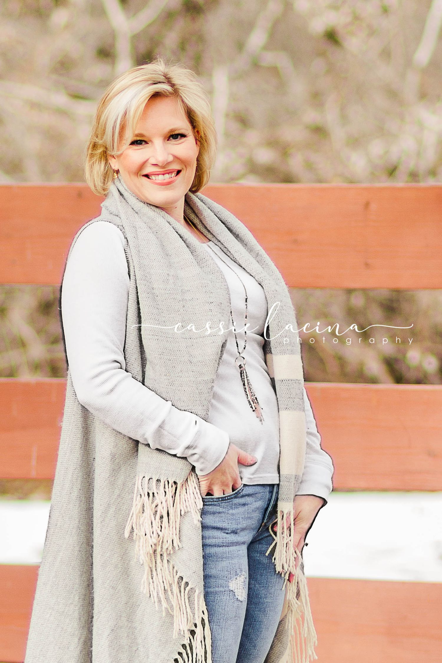 Person in winter attire posing on wooden bench outdoors wearing gray scarf and jeans.