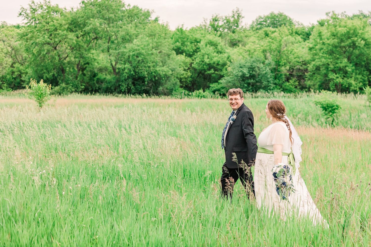 Wedding couple walking through sunny meadow with green trees in background.