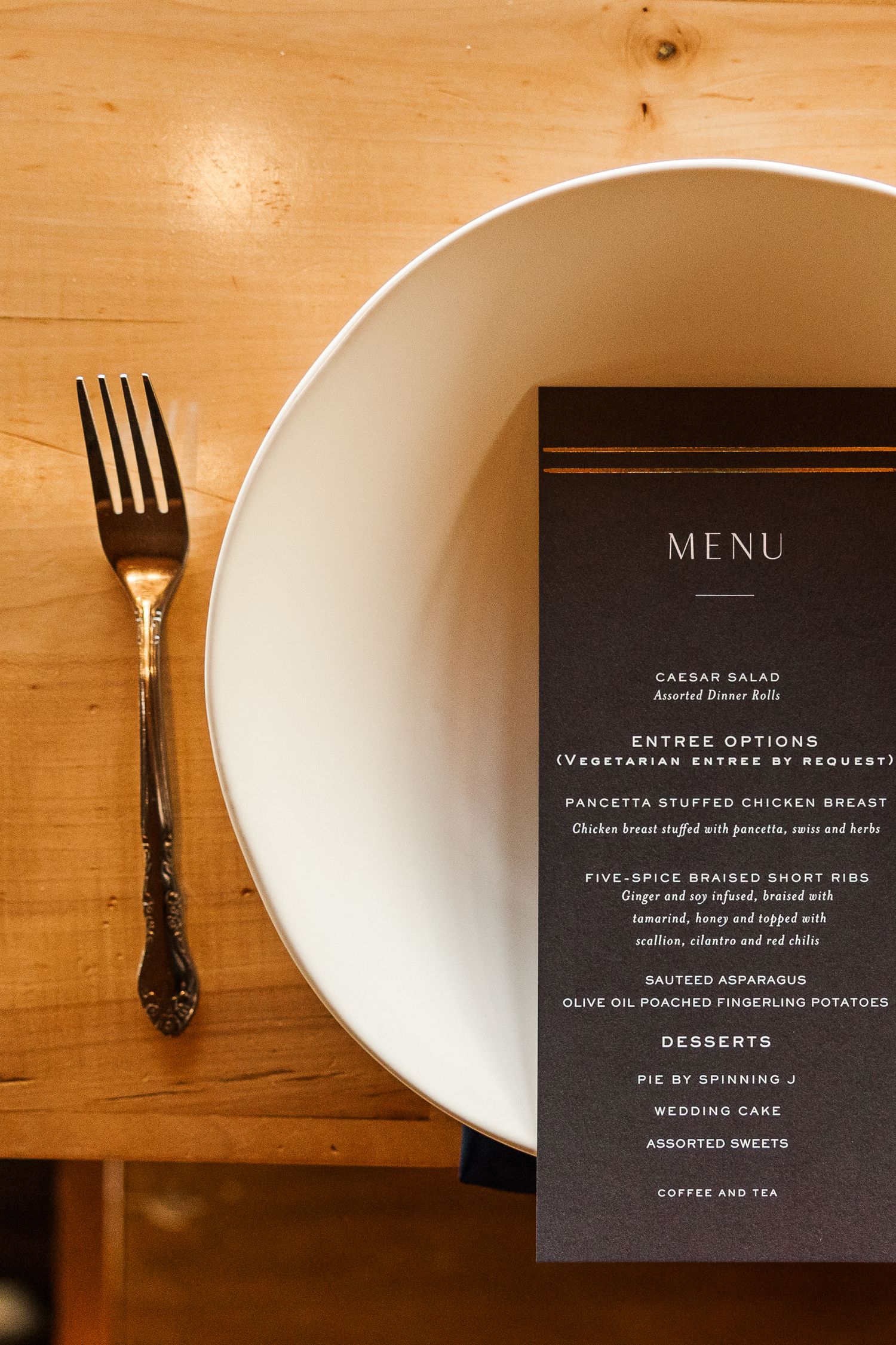 Black menu card and silver fork on white plate against wooden table surface.
