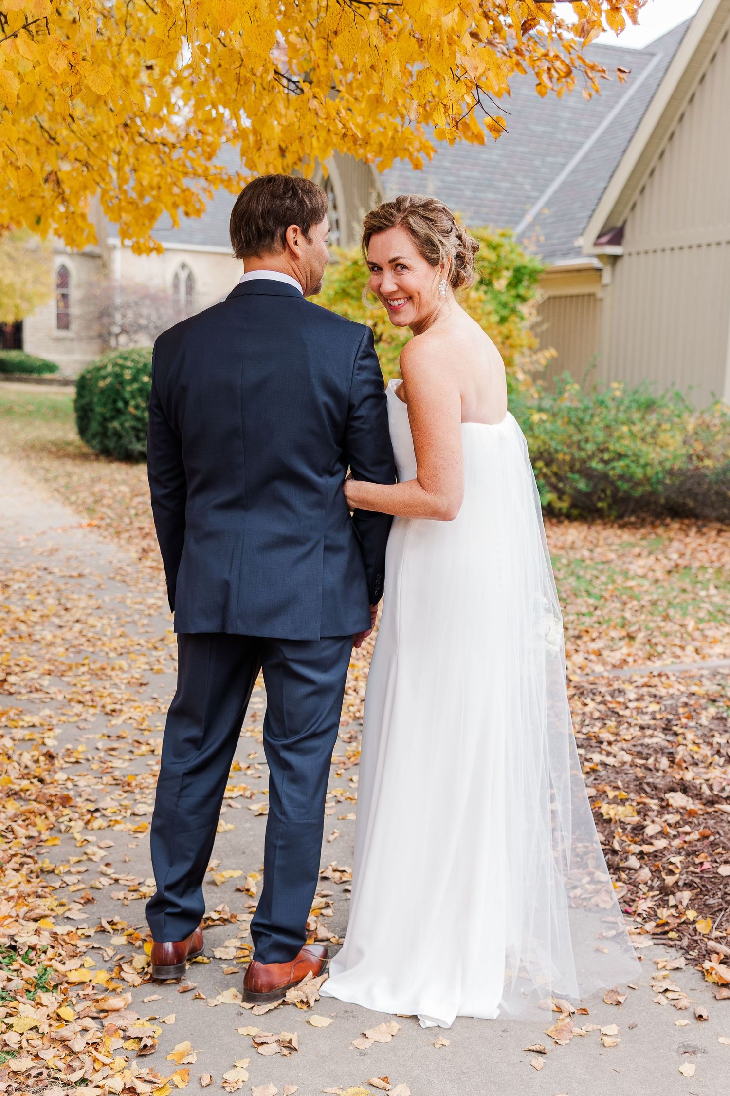 A newlywed couple sharing a playful moment under golden autumn leaves on a garden path.
