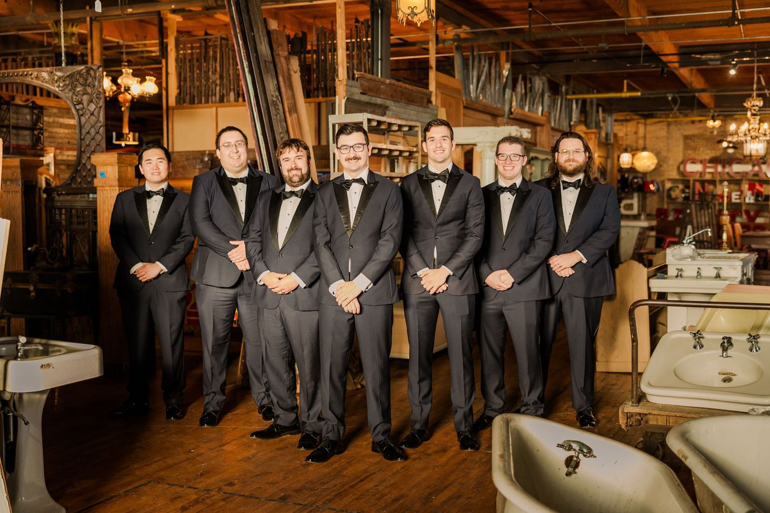 Wedding party in black tuxedos poses in a vintage venue with warm lighting.