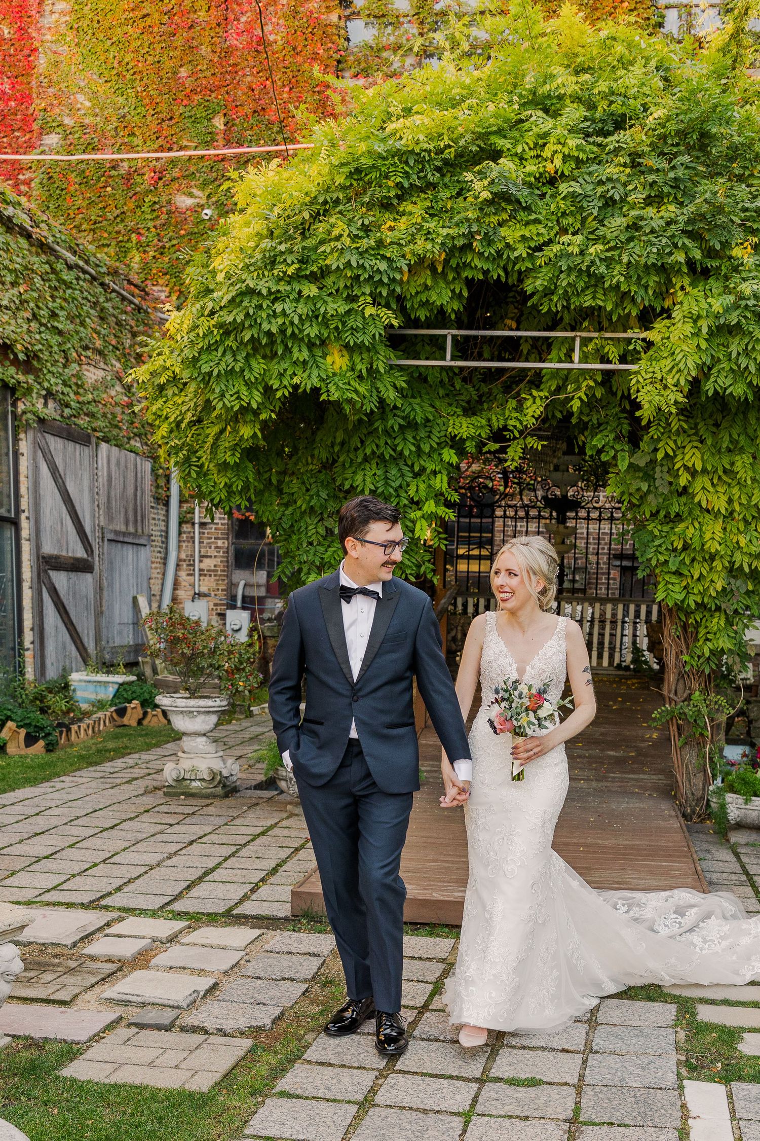 A couple walks together under a lush green archway in an elegant garden courtyard setting.